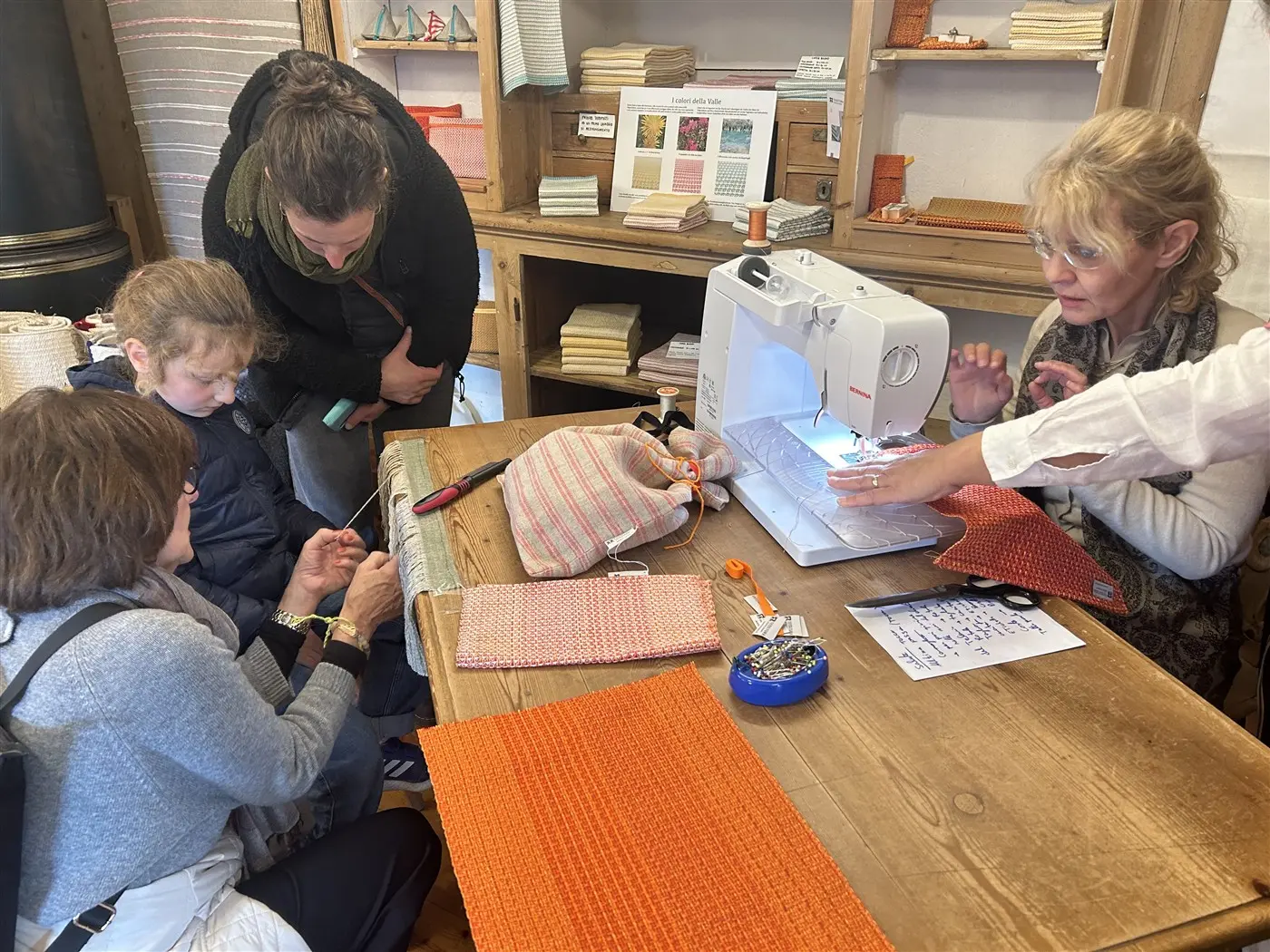 Un gruppo di persone sta partecipando a un laboratorio di cucito. Una donna, seduta accanto a una macchina da cucire, mostra a due ragazze come utilizzare il materiale, mentre altre persone osservano attentamente. Sulla tavola ci sono varie stoffe e attrezzi da cucito.