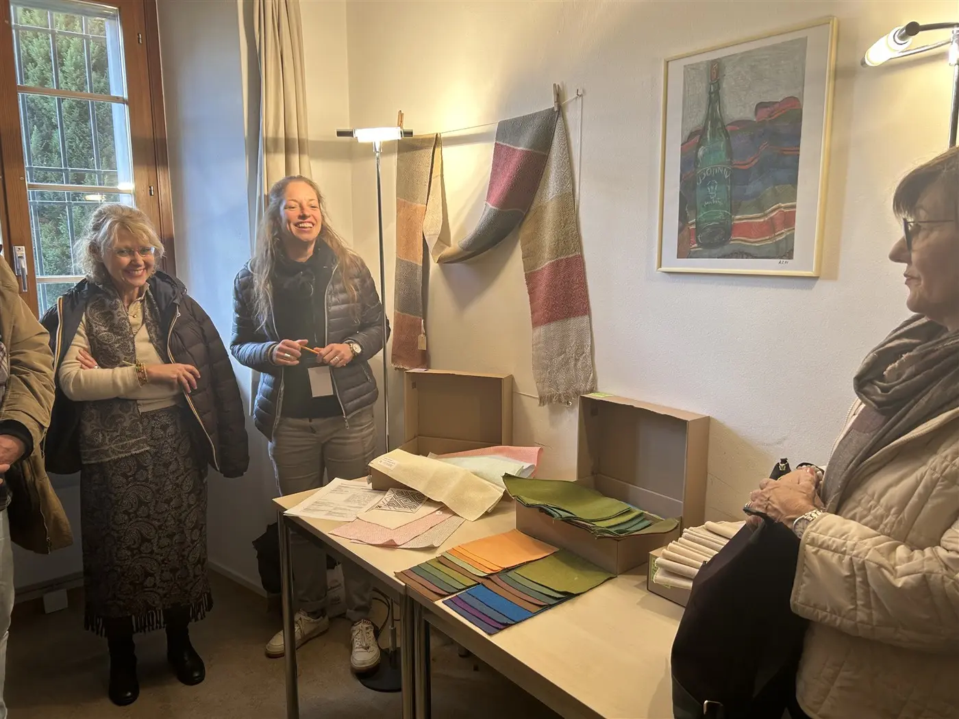 Un gruppo di persone si trova in una stanza luminosa. Due donne sorridono mentre osservano diversi tessuti colorati disposti su un tavolo. Una delle donne è in piedi accanto a un tavolo con campioni di tessuto e un'altra donna è seduta, in attesa di parlare. Sullo sfondo, ci sono pannelli di tessuto appesi e una