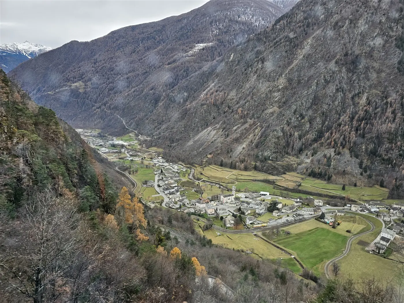 Vista panoramica di una valle montana, con un villaggio circondato da colline e alberi spogliati. In lontananza, si intravedono le cime delle