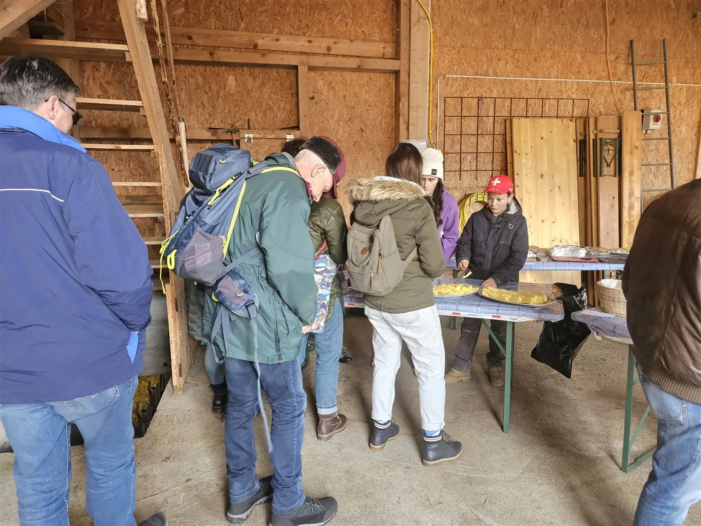 Un gruppo di persone si trova all'interno di un edificio di legno, mentre osservano e scelgono cibo su un tavolo. Alcuni indossano giacche invern