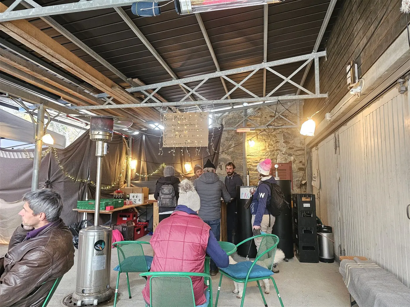 Un ambiente interno di un locale con un soffitto in metallo, diverse persone che parlano tra loro, un bancone con un barista, tavoli e sedie verdi, e un ris