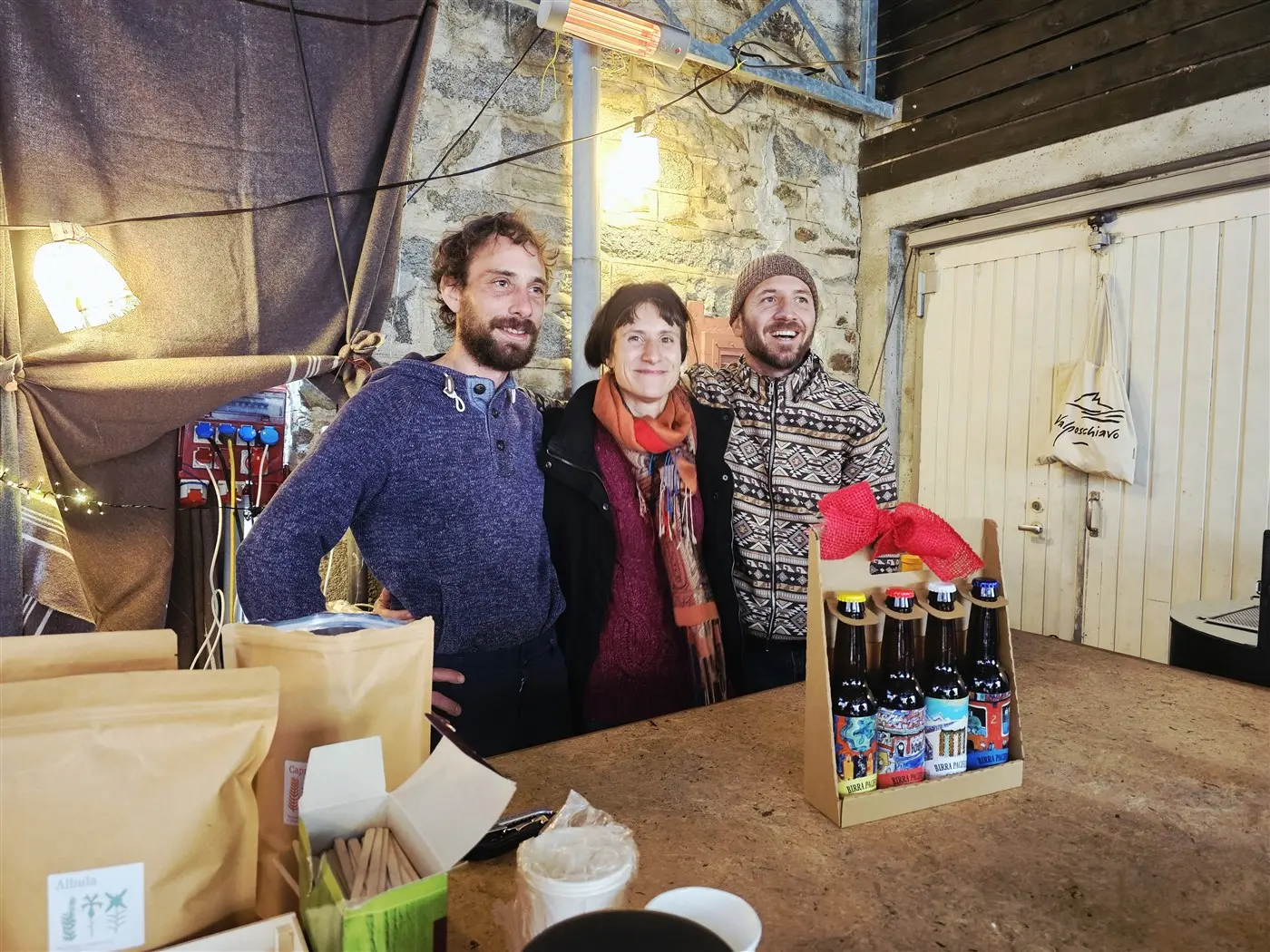 Tre persone sorridenti stanno in piedi dietro a un bancone, con una selezione di bottiglie di birra artigianale in una scatola decorativa. Il fondo presenta pareti