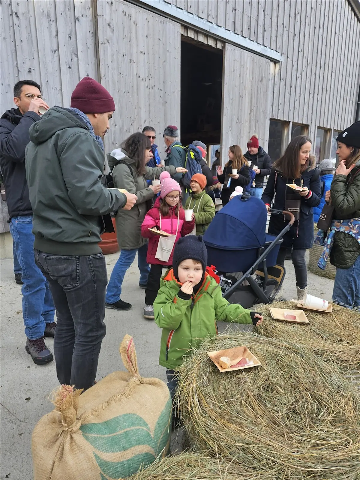Gruppo di persone in un'area all'aperto, alcune con bevande calde mentre altri mangiano. Un bambino in giacca verde si trova in primo piano, seduto su un sac