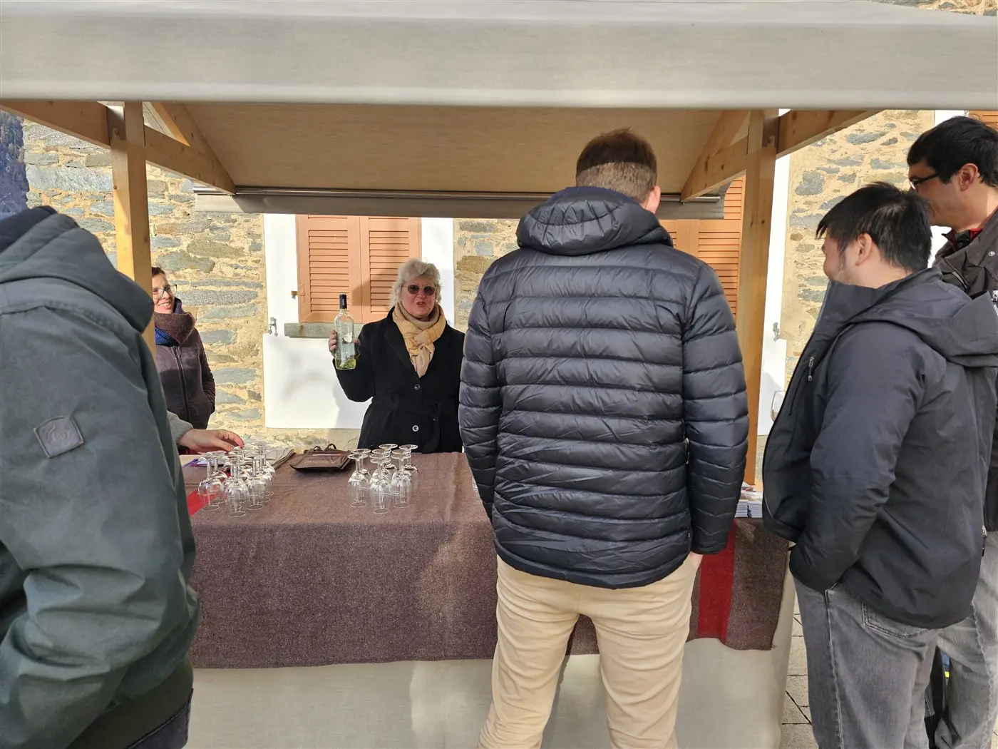 Una donna con i capelli bianchi tiene in mano una bottiglia di vino, mentre è circondata da un gruppo di persone che interagiscono con lei. Il contesto è un mercato o