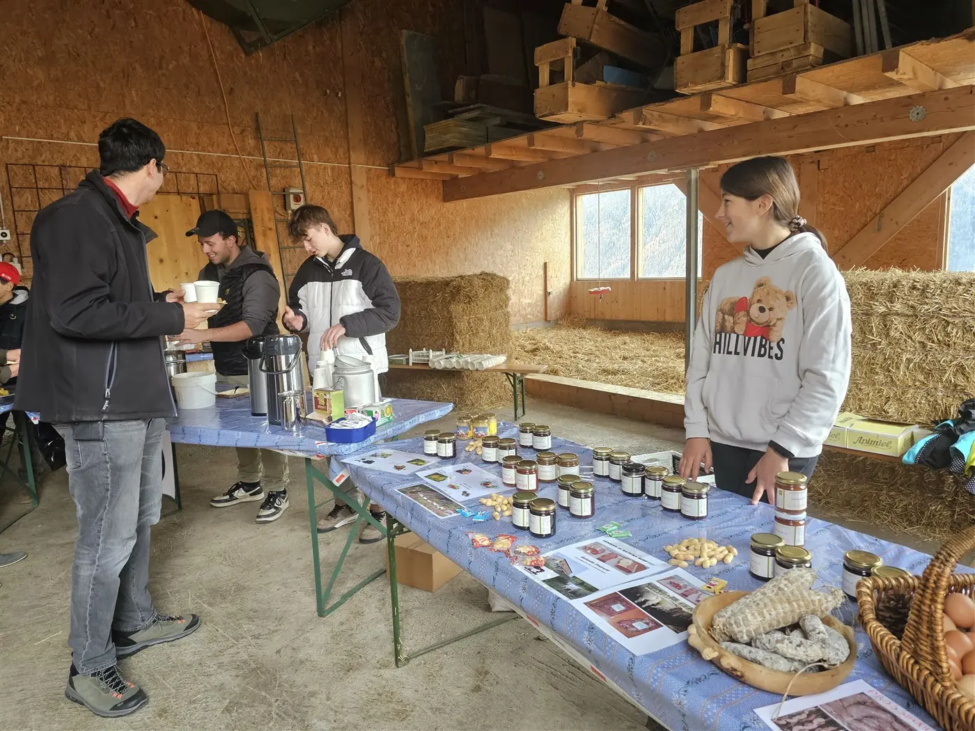 Un mercato di prodotti locali in un barn. Un tavolo è coperto di barattoli di marmellata e conserve, mentre alcune persone interagiscono e degustano prodotti. Sullo sf