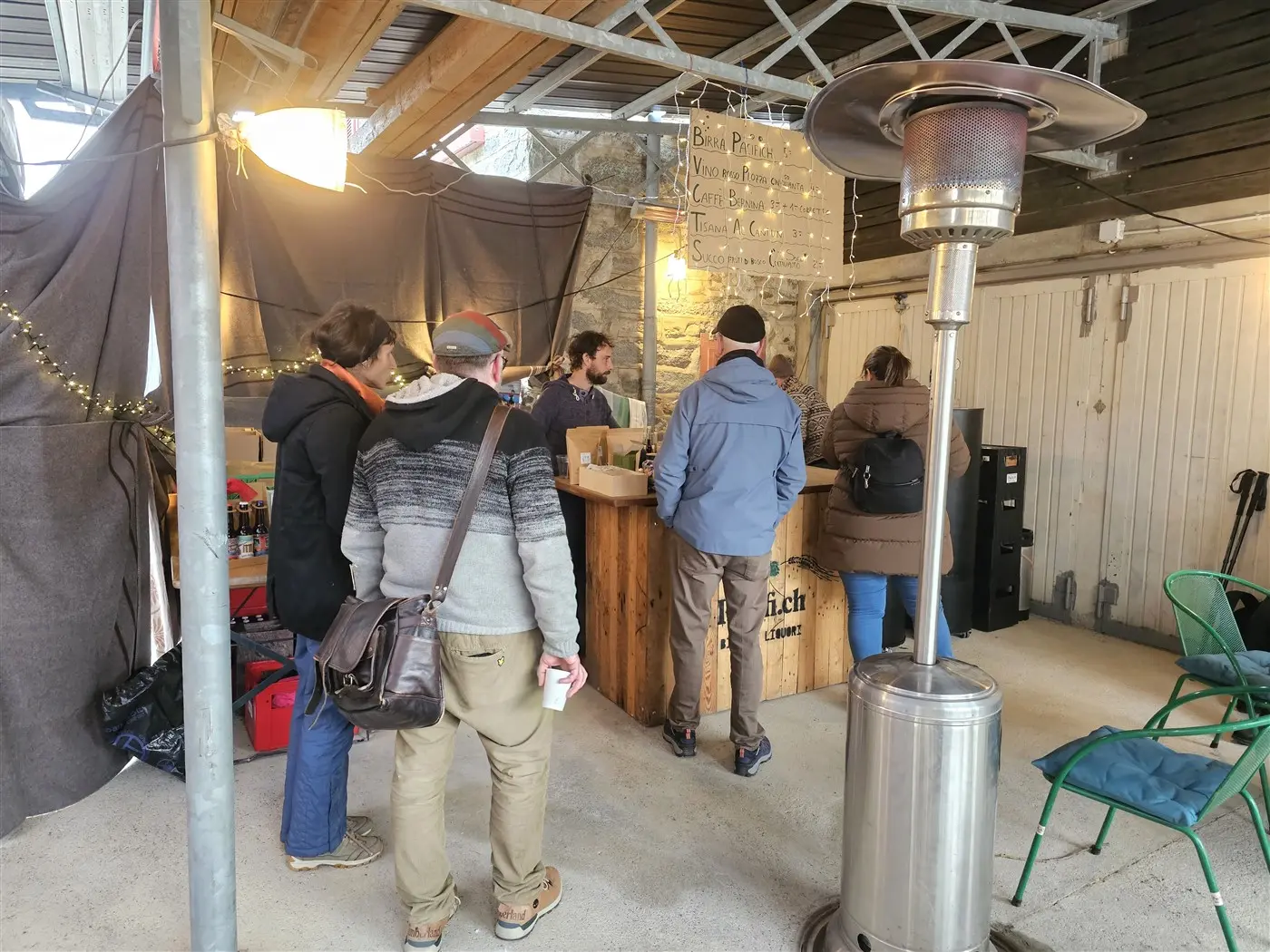 Un gruppo di persone si trova davanti a un bancone di legno in un ambiente esterno coperto. Al bancone ci sono diverse bevande e una persona serve i clienti. In primo piano