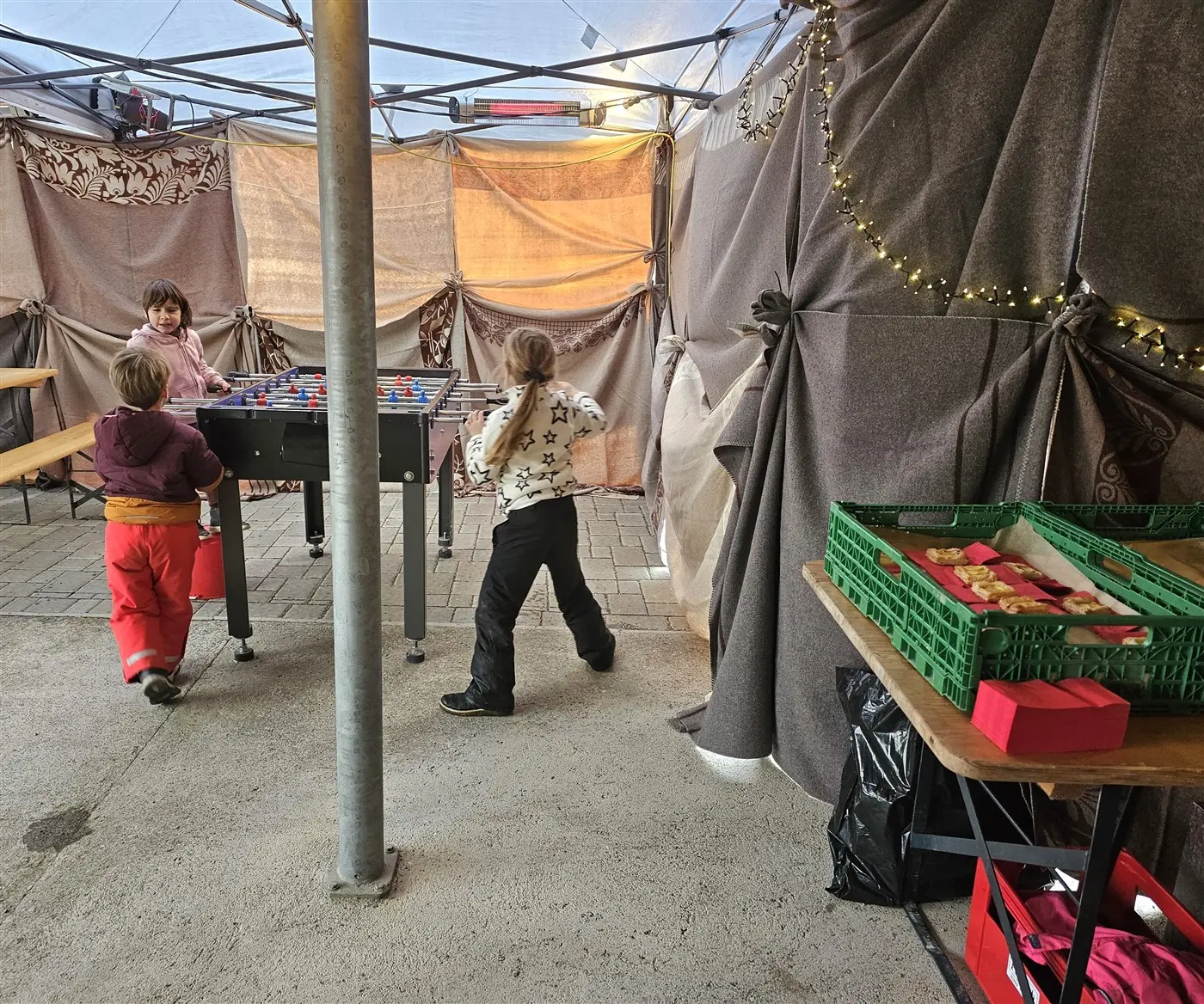 Due bambini stanno giocando a calcetto sotto un tendone. Dietro di loro, si vedono tavoli e una serie di scatole con cibo. Le pareti del tendone sono