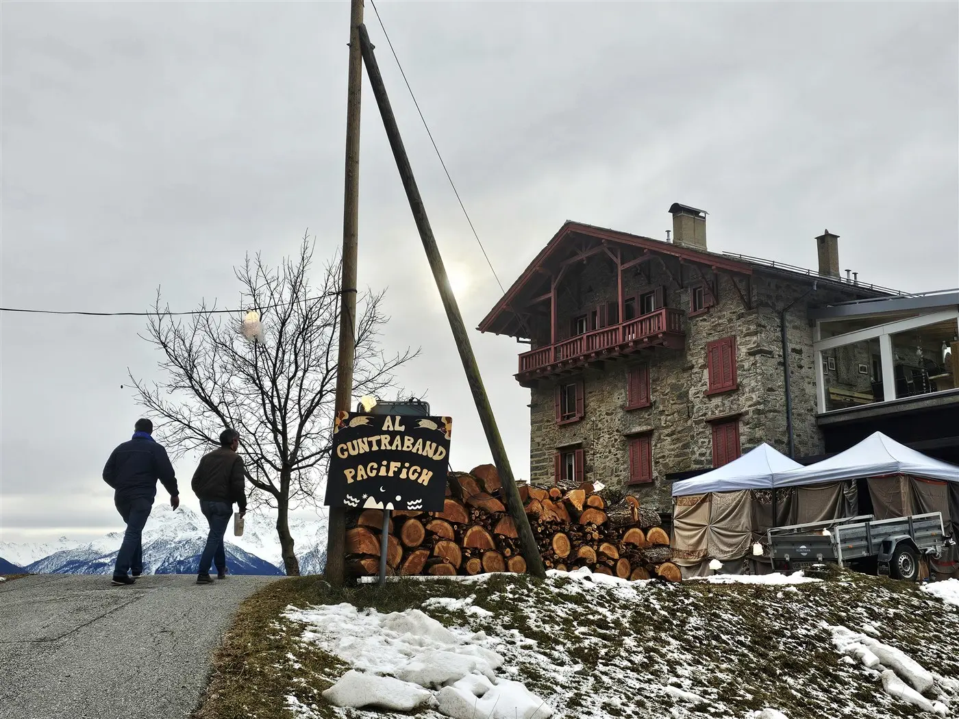 Due persone camminano lungo una strada verso un edificio in pietra con un balcone in legno rosso. A fianco si trova un cartello che indica il ristorante "Al Gu