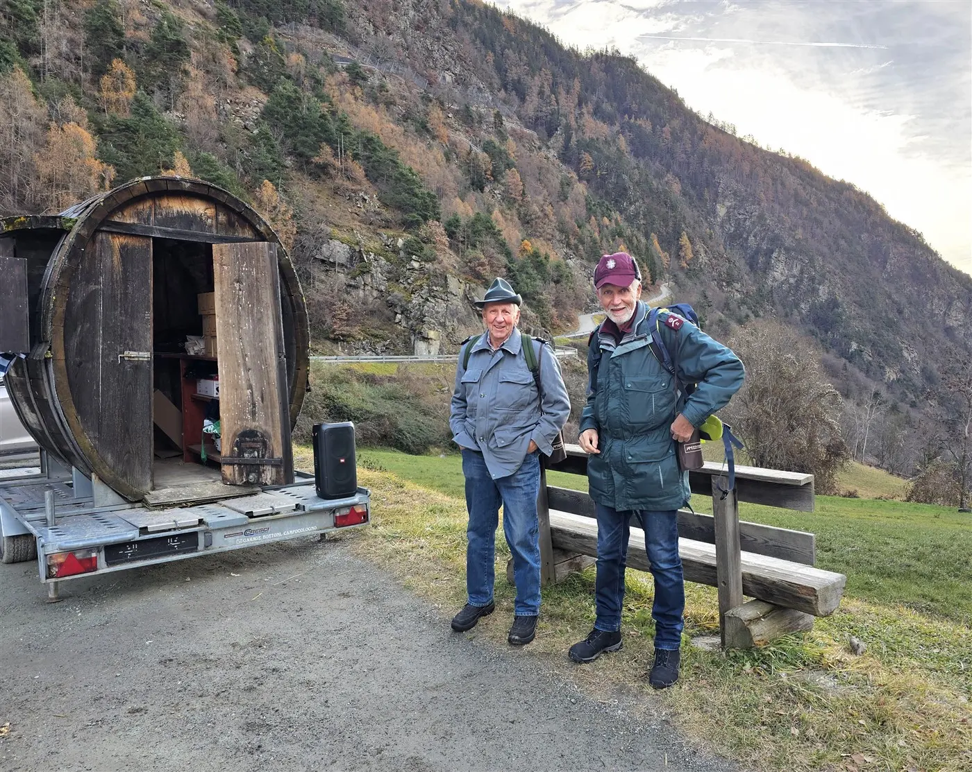 Due uomini sorridenti in abbigliamento casual, uno con un cappello verde e l'altro con un berretto bordeaux, stanno in piedi accanto a un rimorchio con una