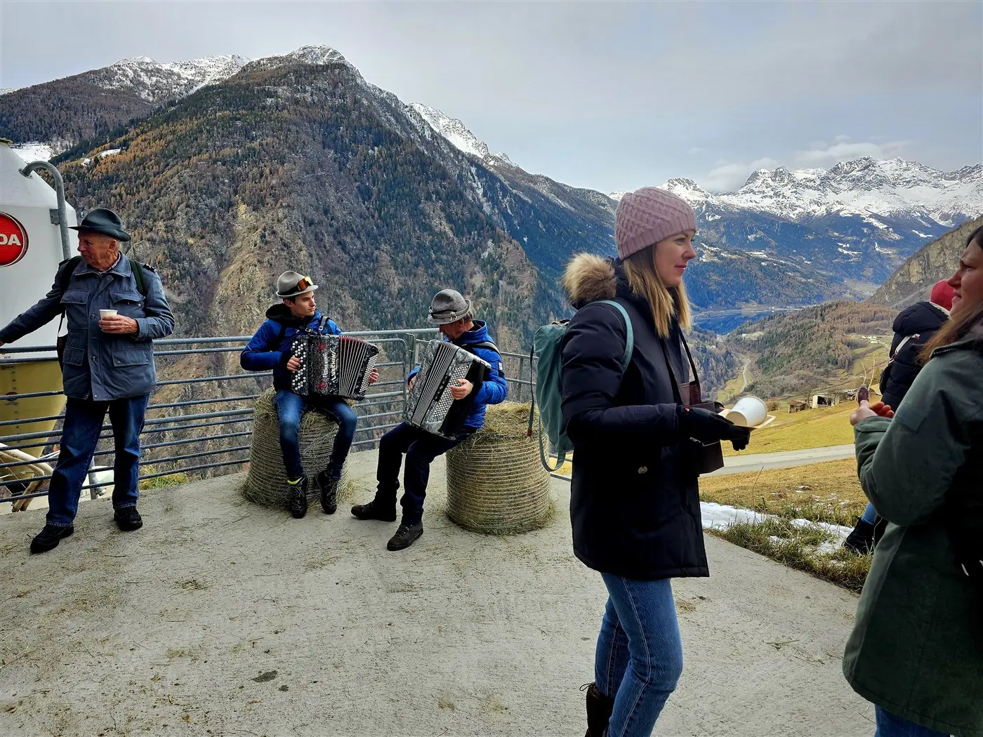 Due musicisti suonano l'organetto in un ambiente montano, mentre un gruppo di persone si gode la vista e una donna in primo piano tiene in mano una bevanda calda, ind