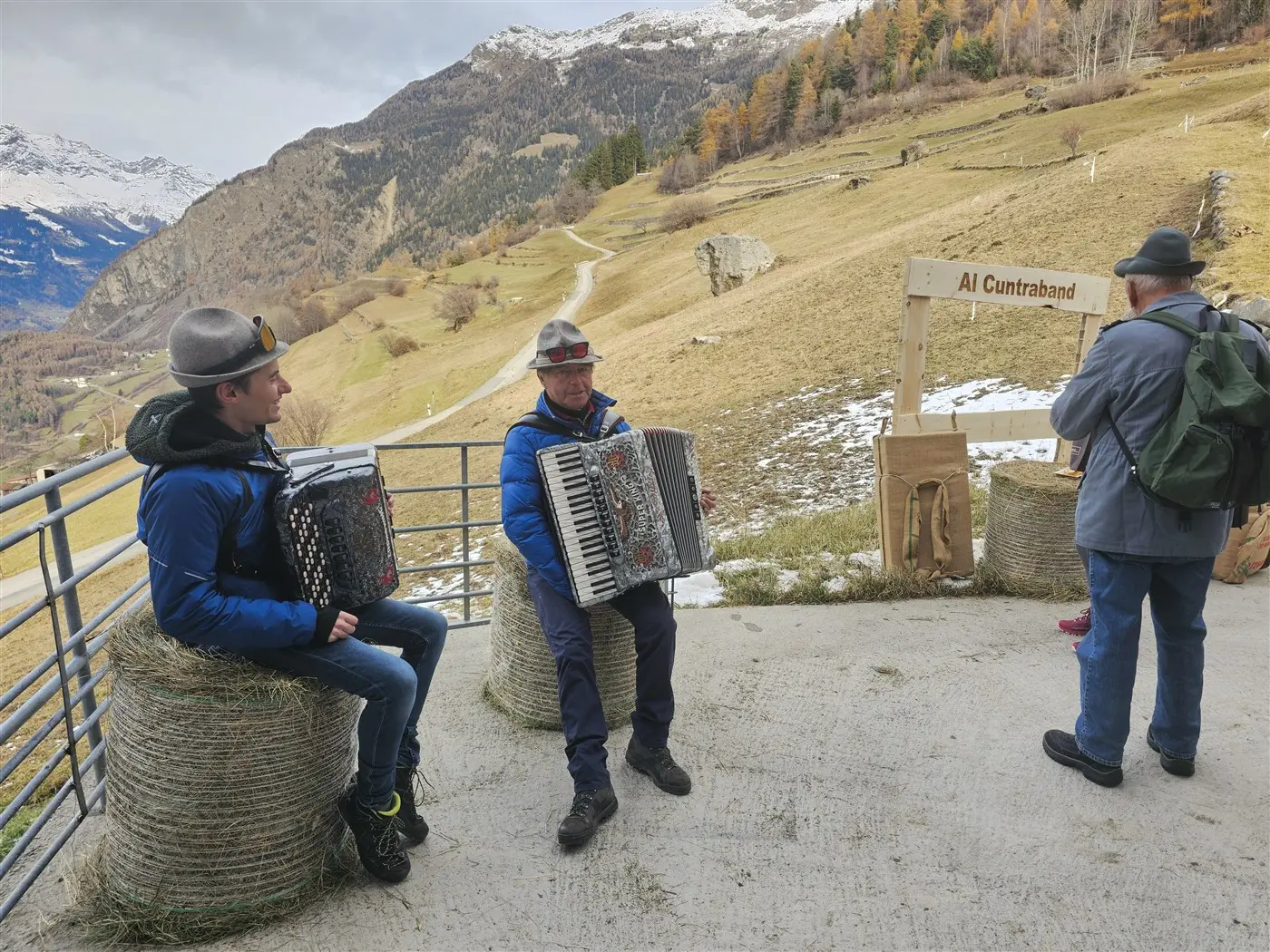 Due uomini suonano l'organetto seduti su balle di fieno, circondati da un paesaggio montano autunnale. Un terzo uomo, in lontananza,