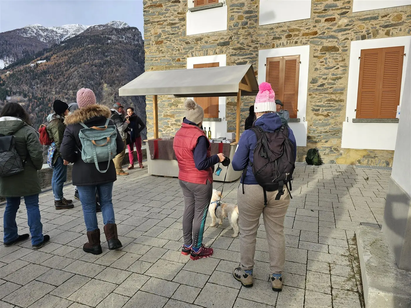 Gruppo di persone in piedi all'esterno di un edificio in pietra, alcuni con cappelli e giacche invernali. Uno di loro tiene un cane al guinzaglio.