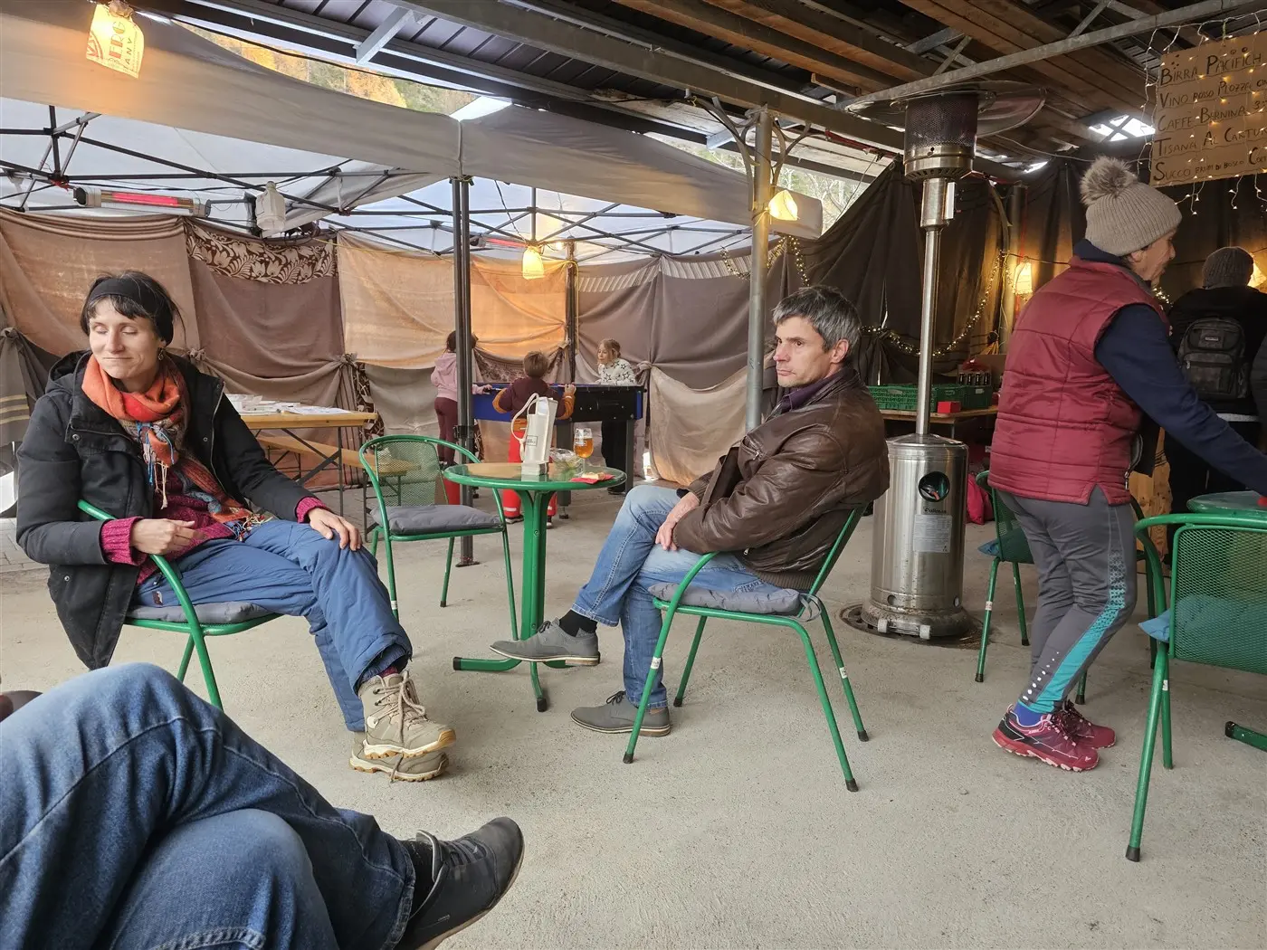 Un gruppo di persone sedute in un ambiente all'aperto, con tavoli verdi e una scaldabere. Una donna indossa una giacca rossa, mentre un uomo in pelle è