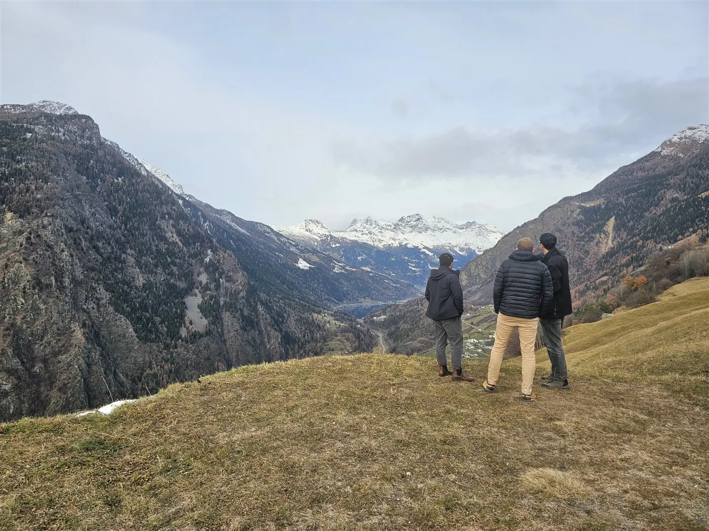 Tre persone osservano un ampio panorama montano, con vette innevate e valli verdi. Il cielo è parzialmente nuvoloso.