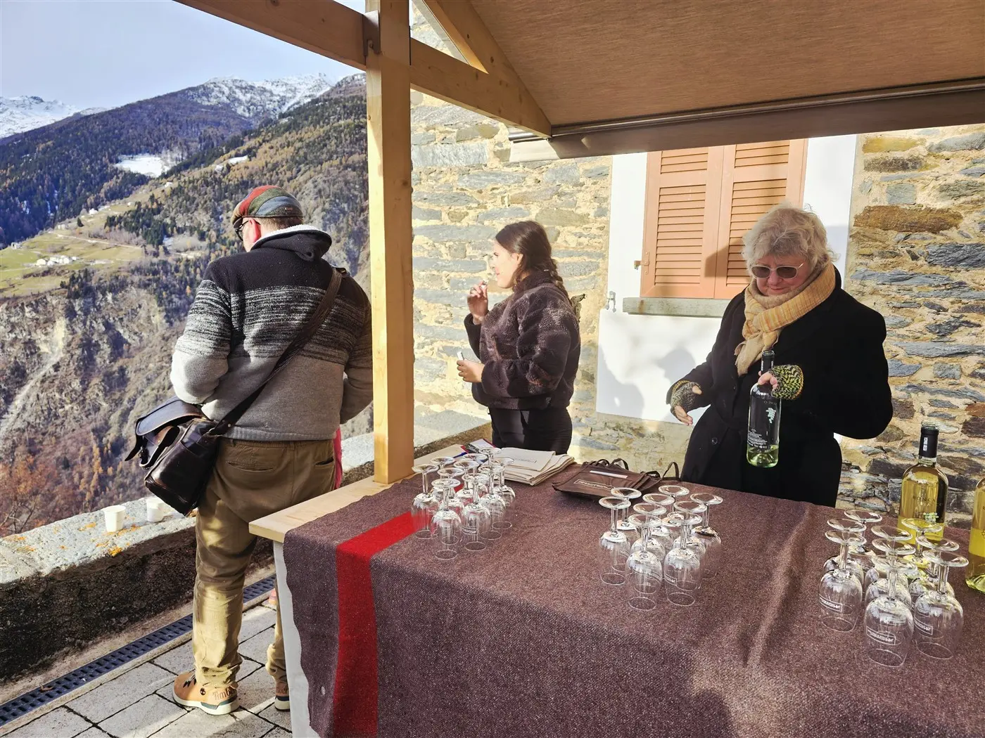 Due persone cercano degustazioni all'aperto in montagna, con una donna che versa bevande e un uomo che osserva il paesaggio. Un tavolo con bicchieri è allest