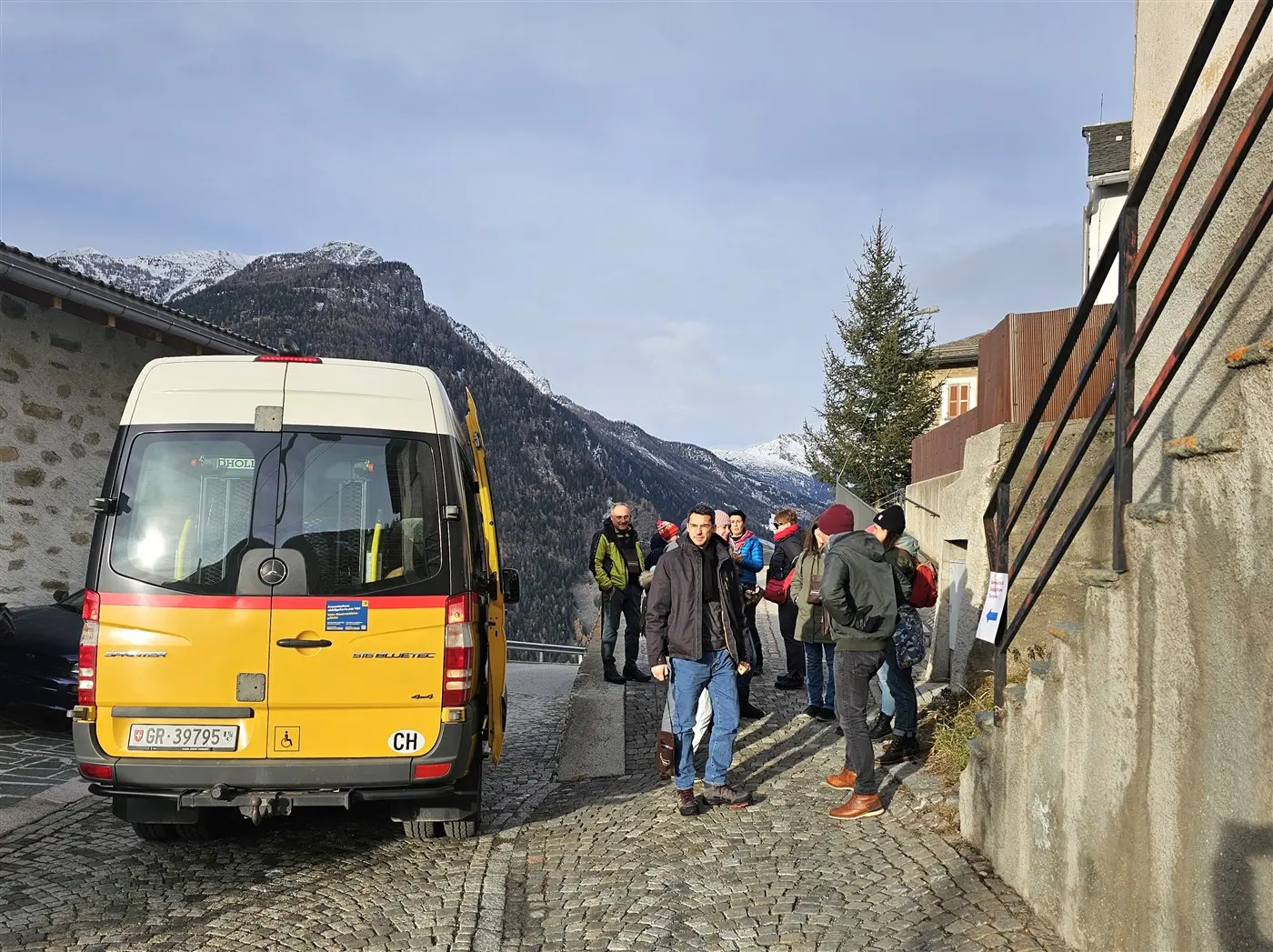 Gruppo di persone in piedi vicino a un autobus giallo in una zona montuosa, con montagne innevate sullo sfondo. Alcuni partecipanti sembrano prepararsi per un'