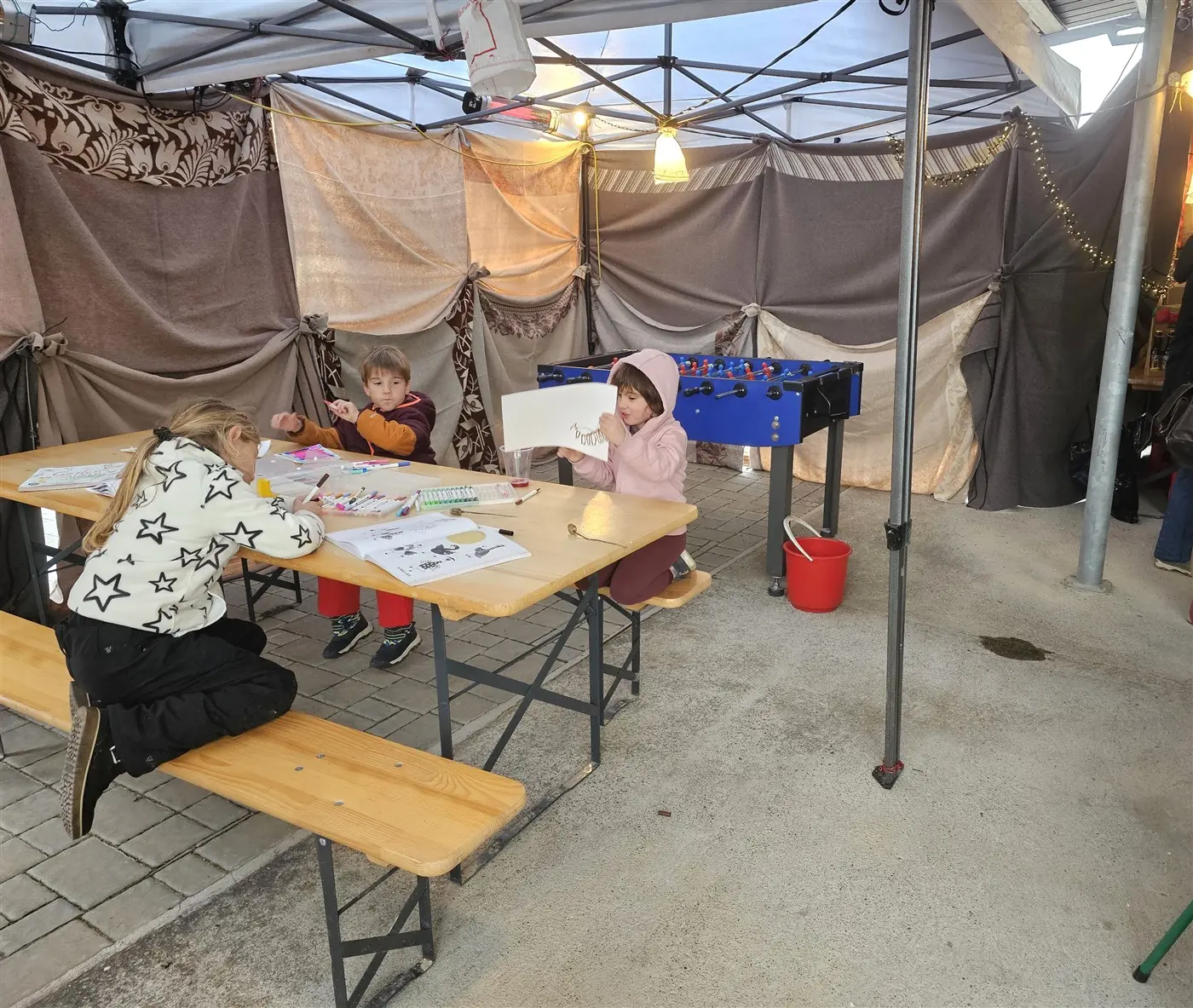 Tre bambini seduti a un tavolo sotto una struttura coperta. Due bambini stanno colorando e disegnando, mentre un terzo gioca a un tavolo da calcio. Il ambiente è