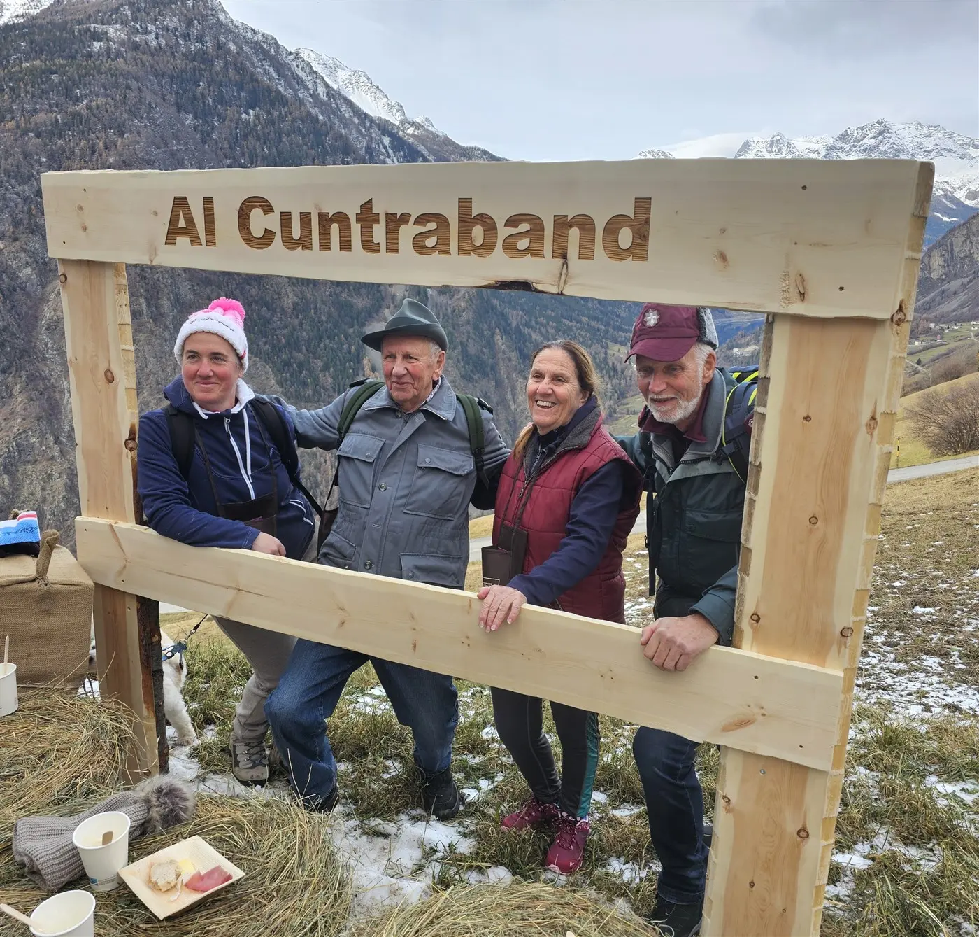 Quattro persone sorridenti posano davanti a un grande fotogramma di legno con la scritta "Al Cuntraband". Sullo sfondo si vedono montagne e un paesaggio