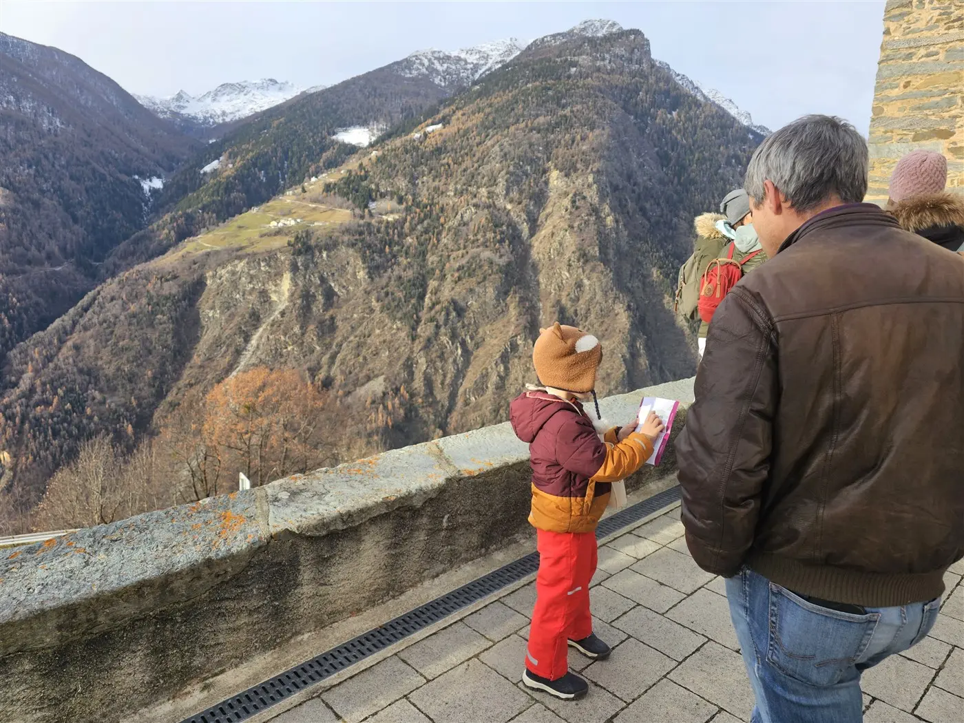 Un bambino con un cappello e una giacca colorata scrive su un quaderno mentre osserva un paesaggio montano. Un adulto, di spalle, si trova vicino a lui