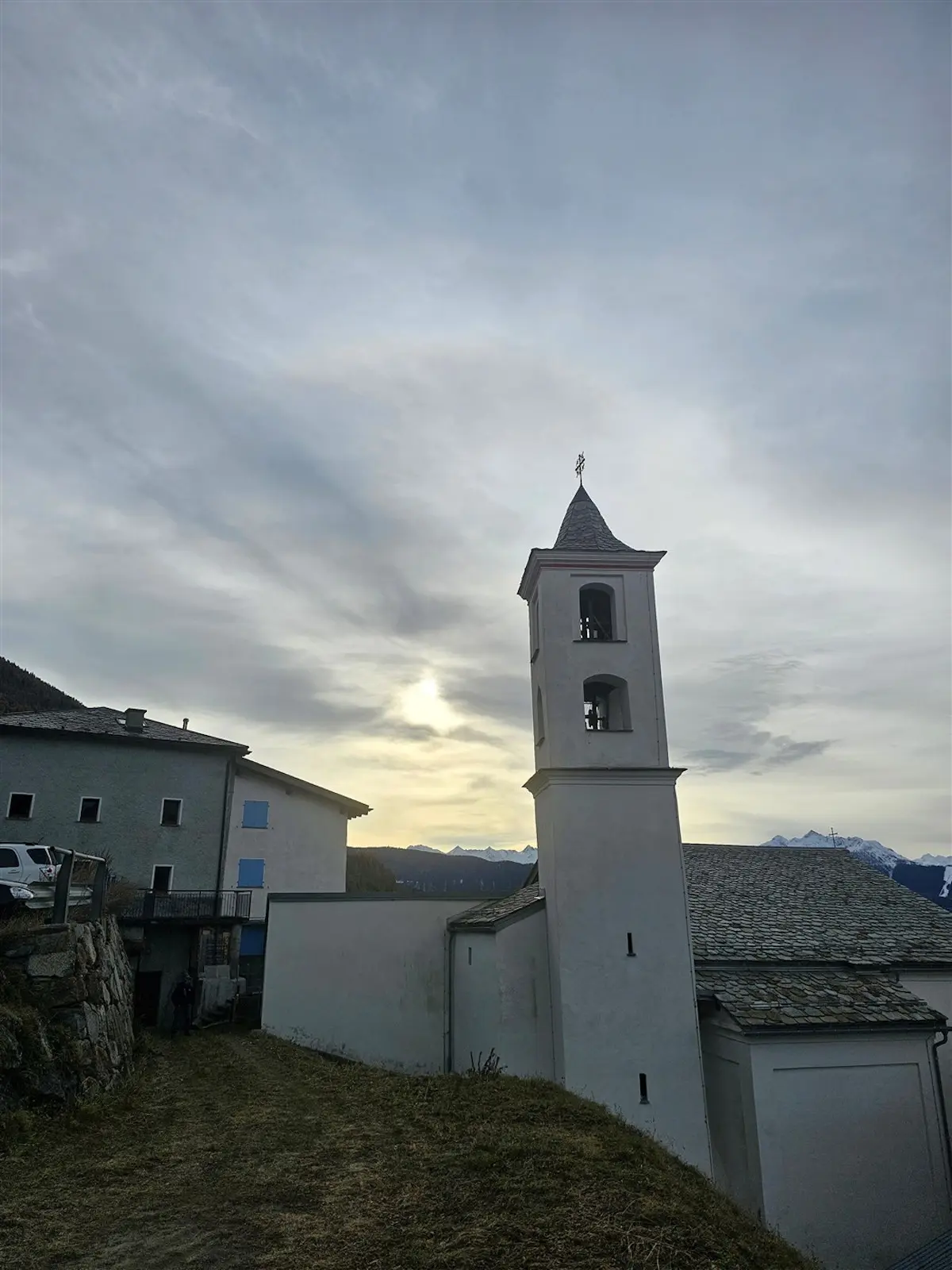 Una chiesa con un campanile slanciato in un paesaggio montano, circondata da un cielo nuvoloso e le montagne sullo sfondo.