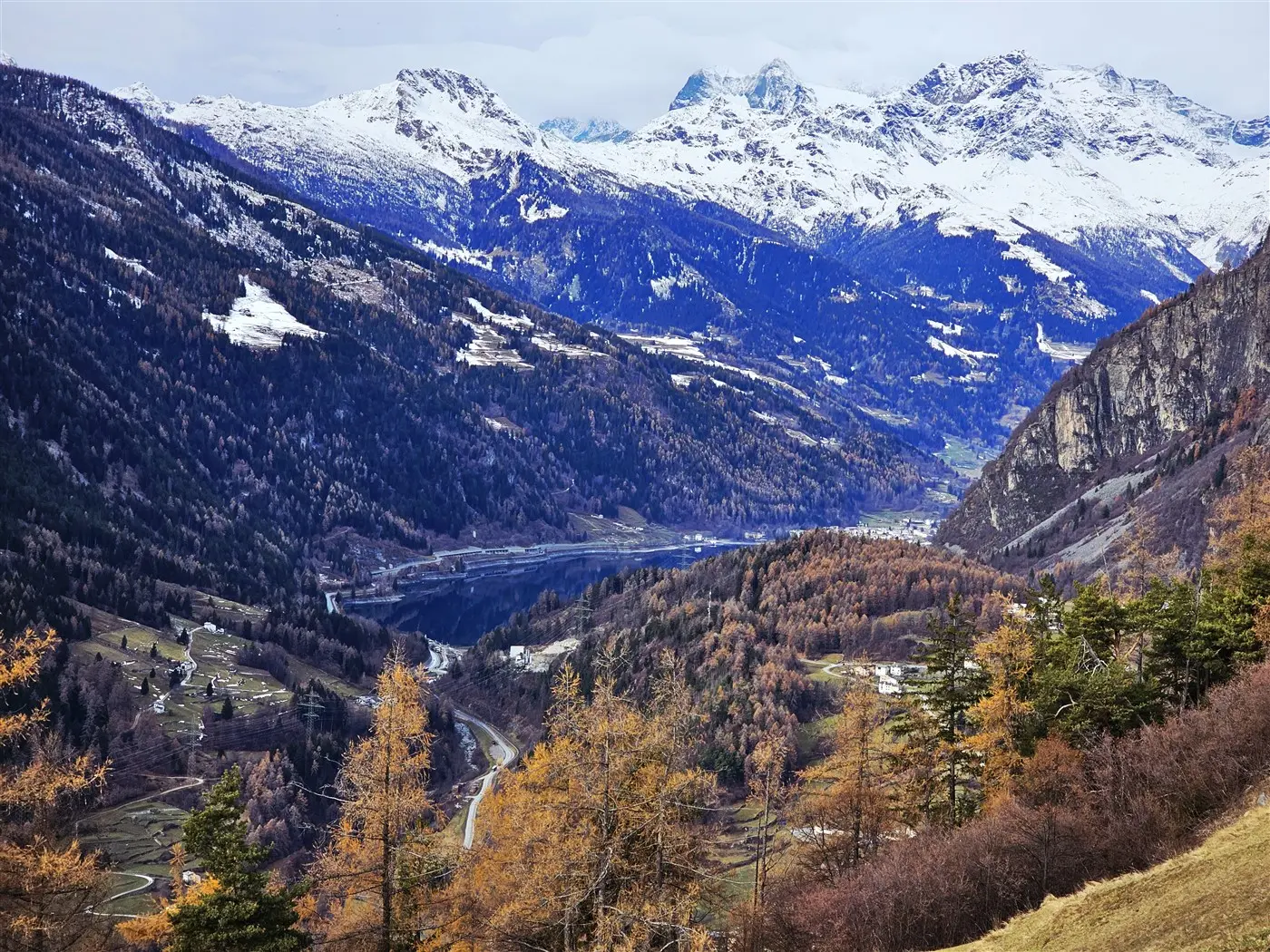 Vasta valle montuosa con cime innevate, alberi di larice arancioni in autunno e un fiume che attraversa il paesaggio.