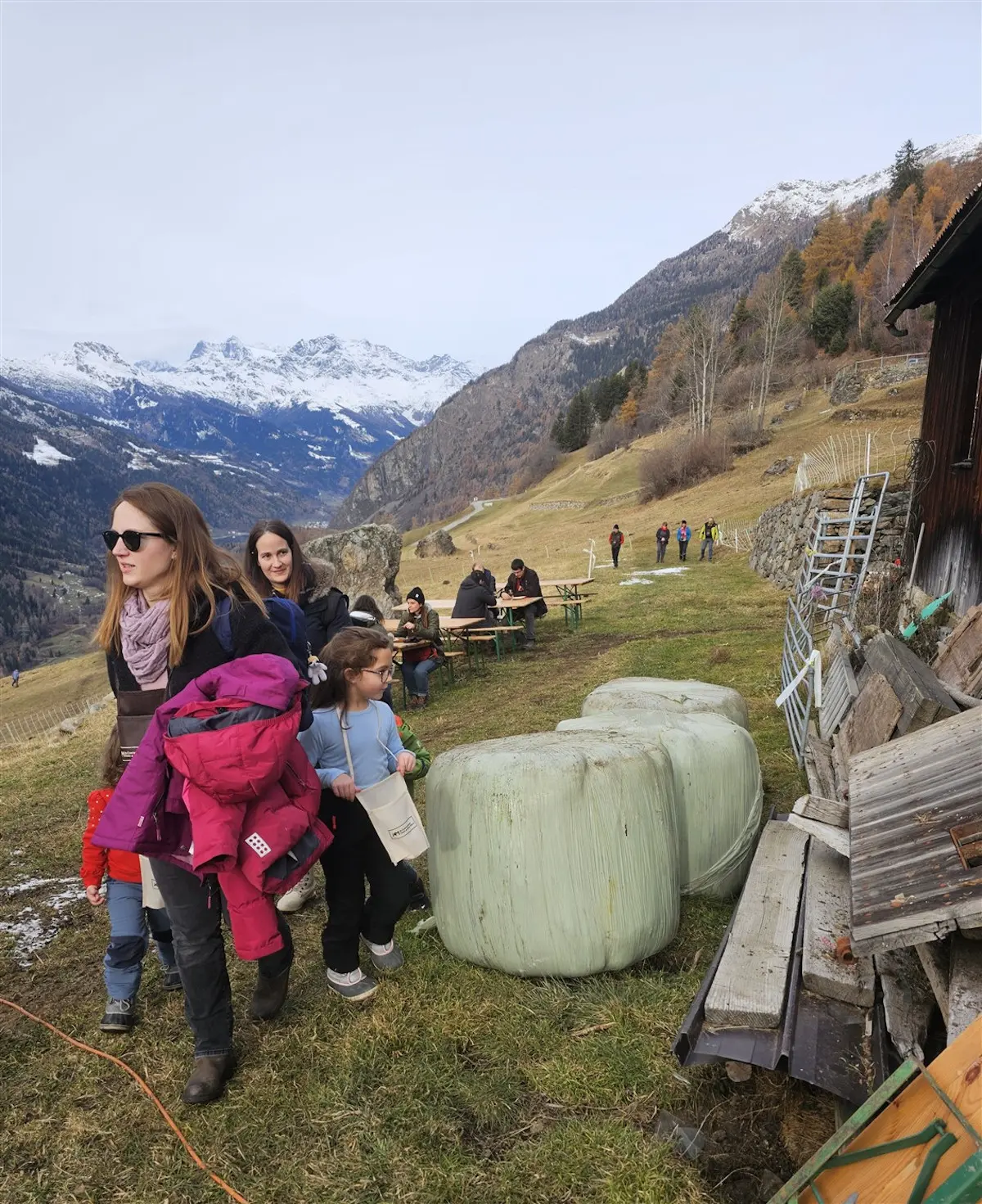 Gruppi di persone passeggiano in un paesaggio montano, con ballette di fieno in primo piano. Sullo sfondo si vedono montagne coperte di neve e un