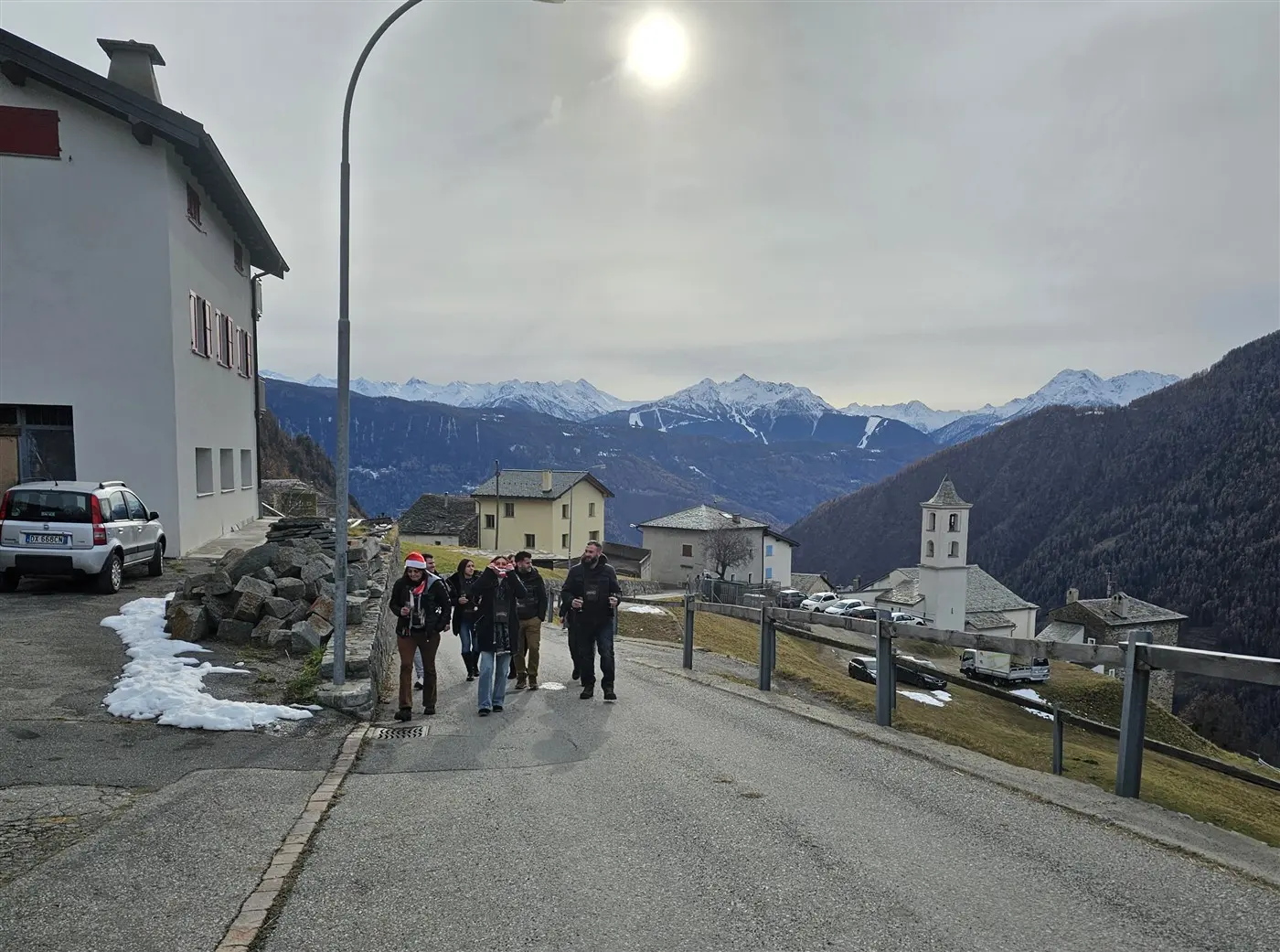 Gruppo di persone che camminano lungo una strada di montagna, circondati da case tipiche e un paesaggio montuoso con cime innevate sullo sfondo. Il
