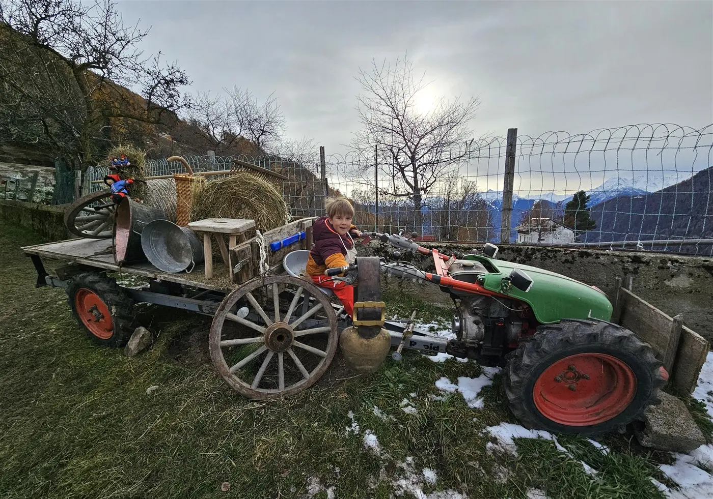 Un bambino sorride mentre si siede su un trattore modificato, circondato da attrezzi agricoli e balle di fieno, con il sole che tramonta sullo sfondo