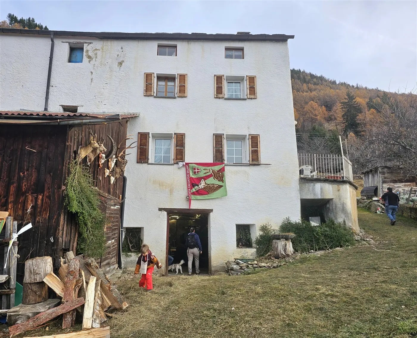 Una casa bianca con finestre di legno a persiana, decorata con una bandiera. Al piano terra, una donna in abiti rossi esce da una porta, mentre un