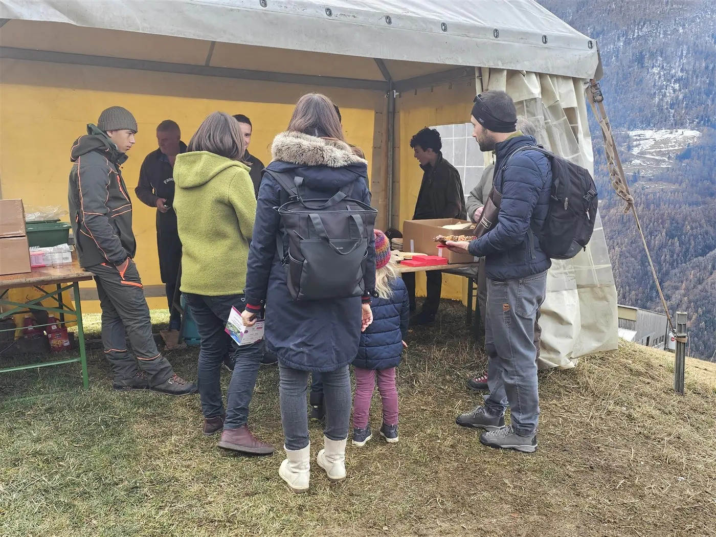 Un gruppo di persone si riunisce sotto un tendone all'aperto per ricevere cibo. Alcuni adulti conversano con un uomo al tavolo, mentre una bambina osserva
