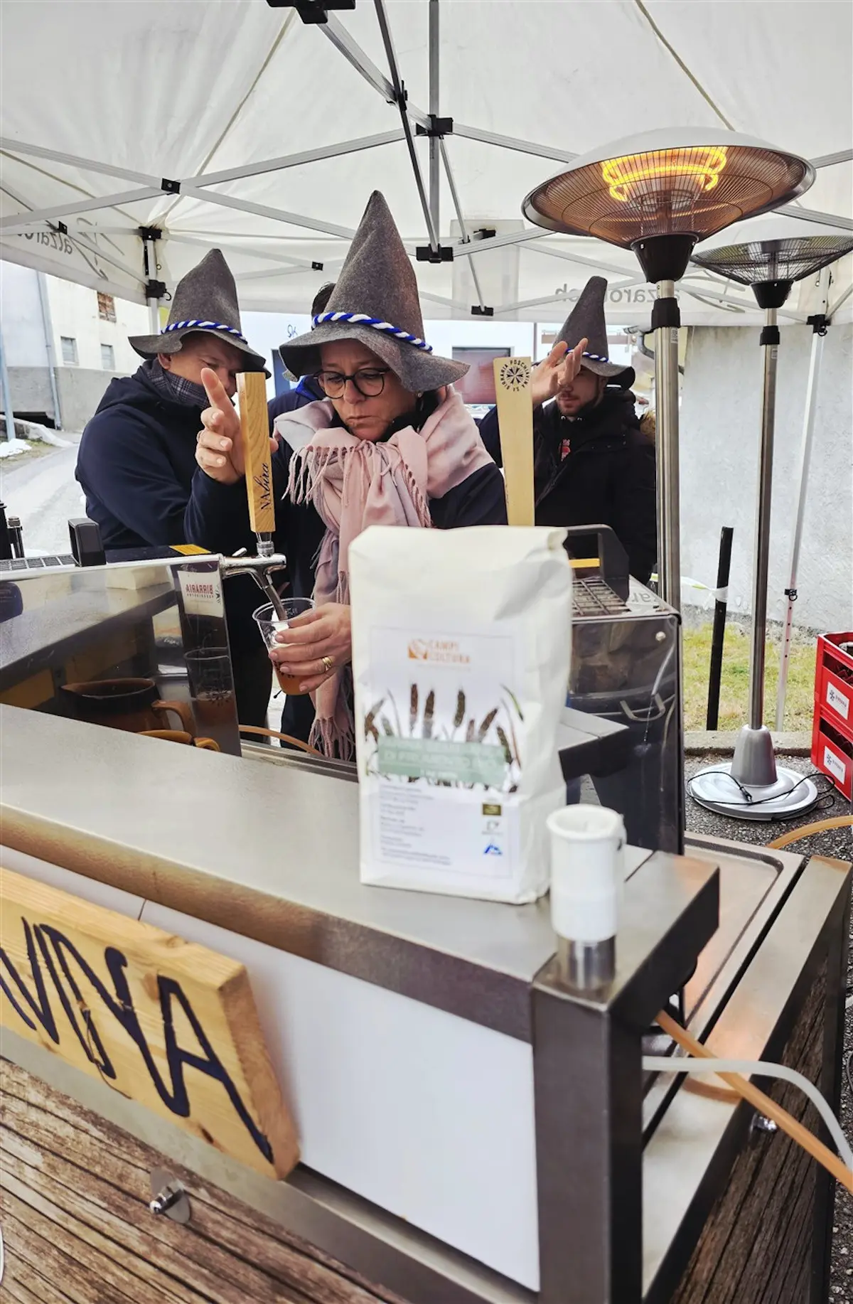 Due persone con cappelli a punta stanno servendo bevande a un banco, mentre una donna indossa una sciarpa e sorseggia da un bicchiere. Sul bancone c