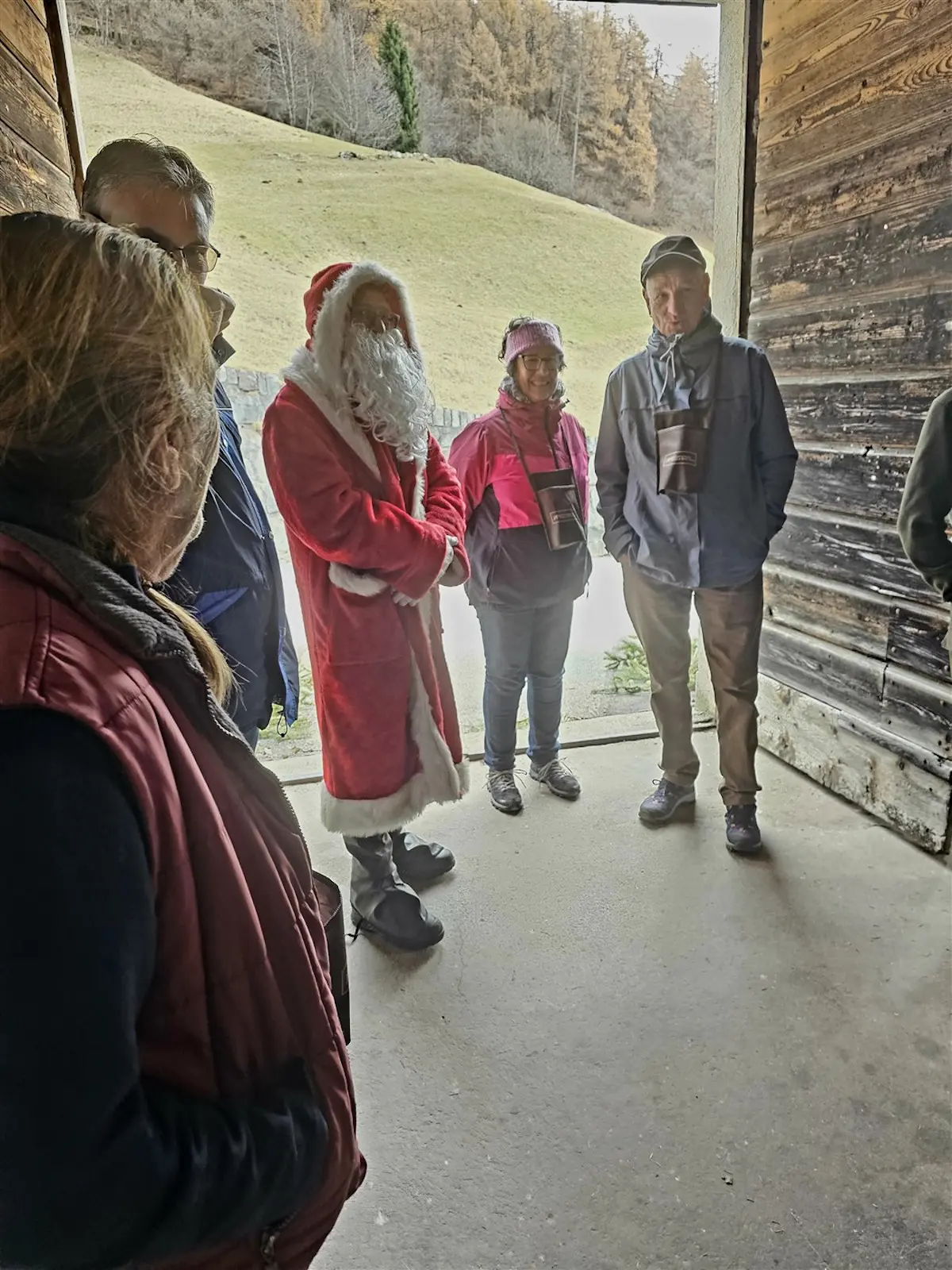 Un gruppo di persone all'interno di un edificio rustico. Un uomo con un costume da Babbo Natale si distingue tra i partecipanti, mentre gli altri indossano abbigliamento caldo