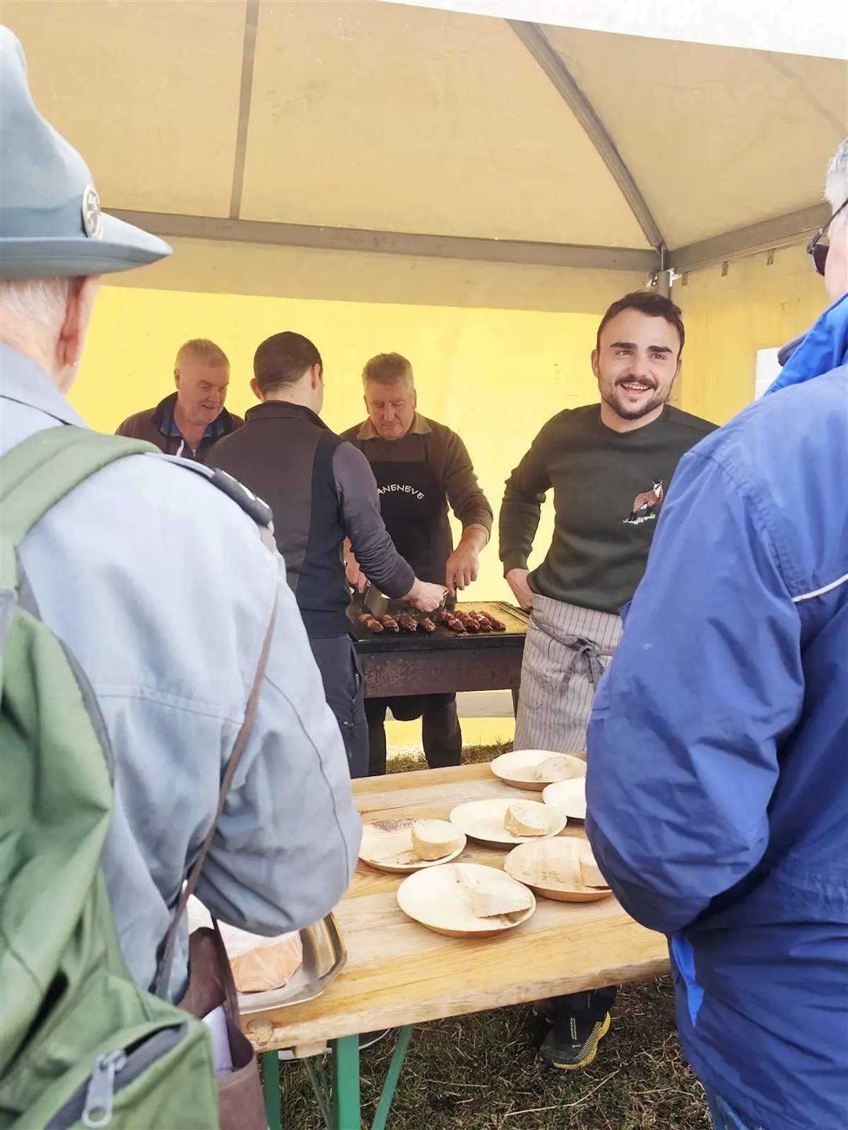 Un gruppo di uomini accanto a un bancone, mentre preparano e servono cibo in un tendone. Alcuni piatti di legno sono disposti su un tavolo. Una