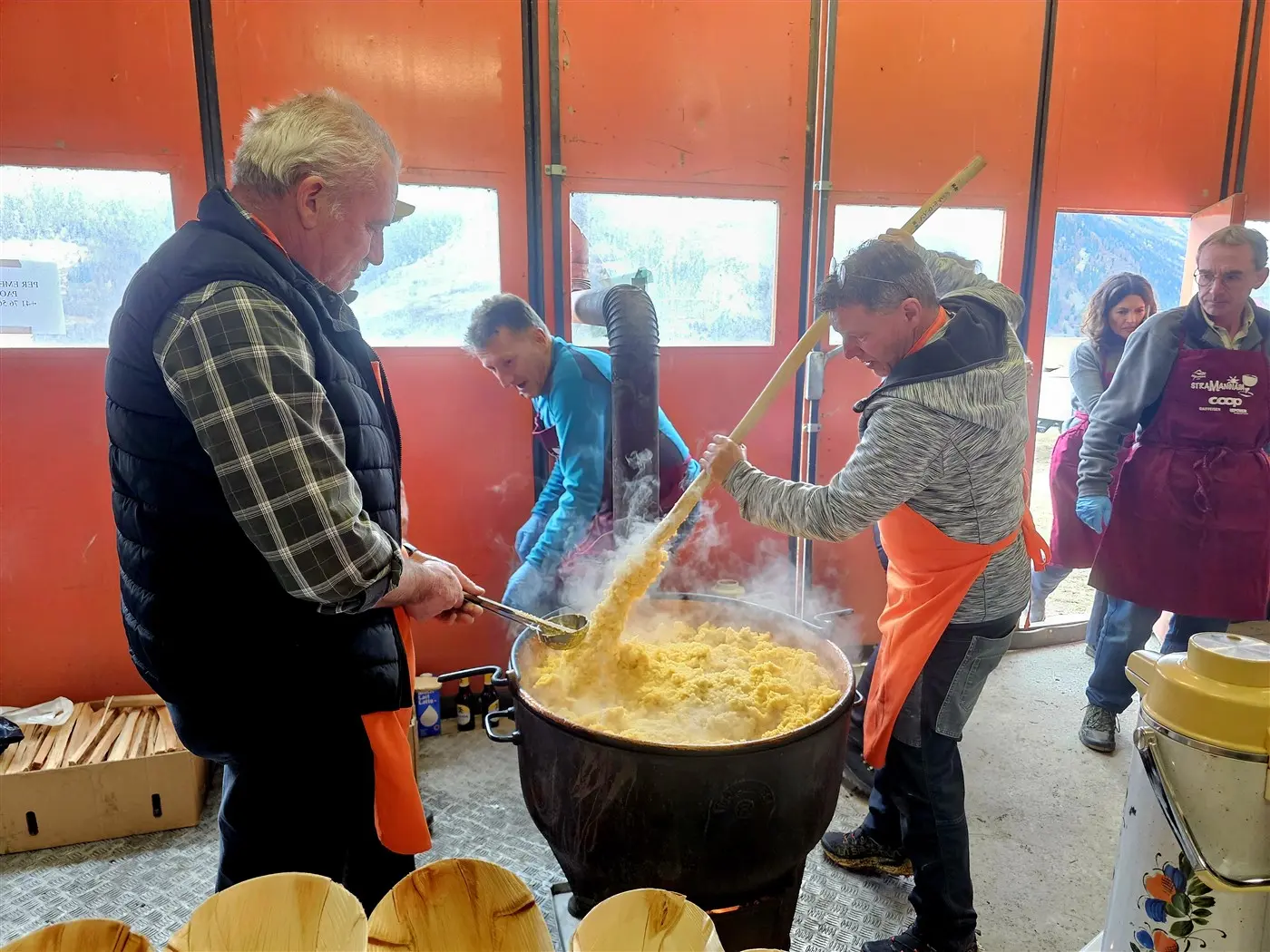 Uomini e donne in un ambiente di cucina, intenti a mescolare un grande pentolone di polenta fumante. Indossano grembiuli arancioni e sorridono mentre lavor