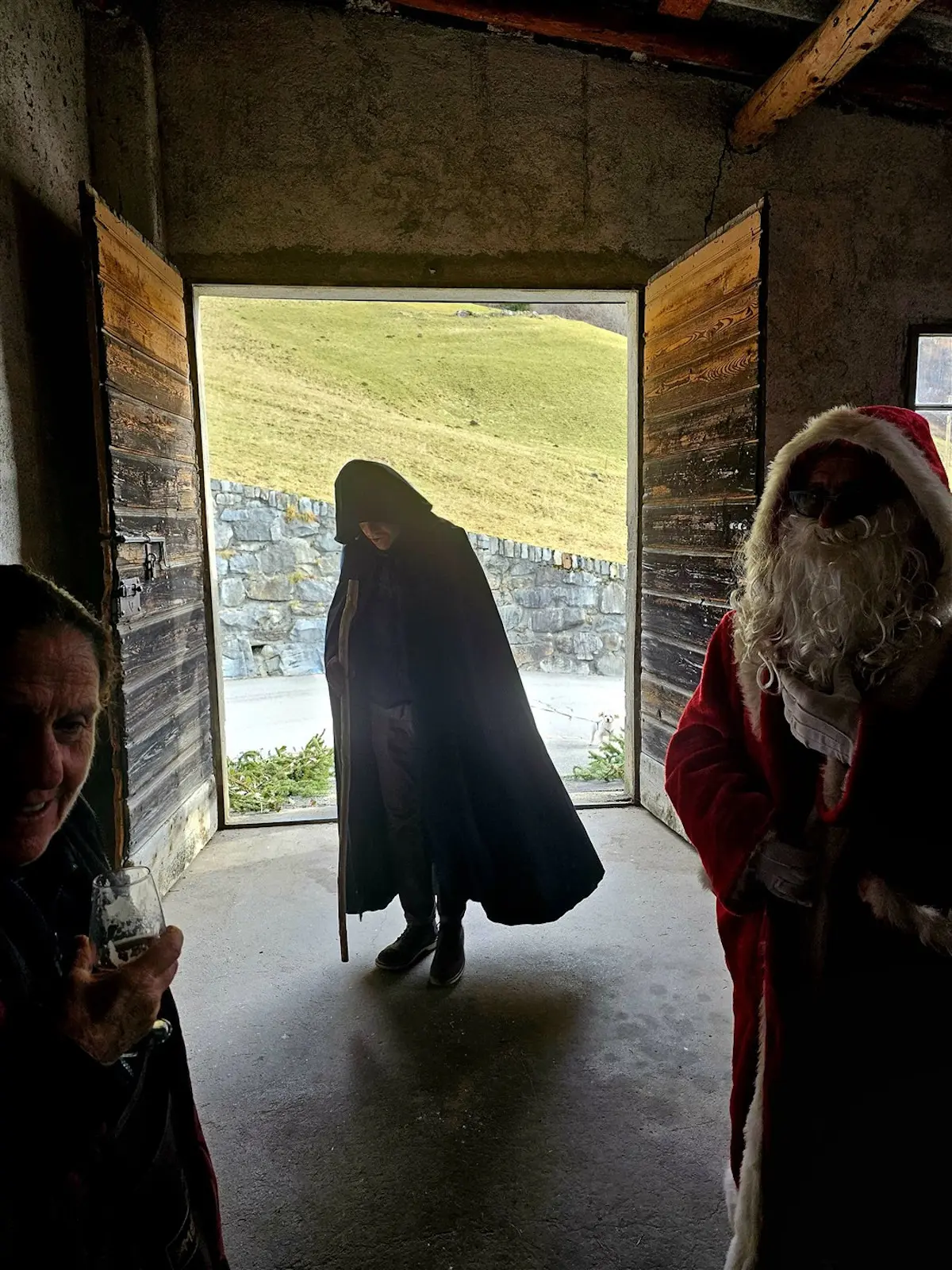 Tre persone in un ambiente rustico. Due figure sono in costume: una veste nera con cappuccio e un Babbo Natale con un sacco rosso. Sullo sfondo, una