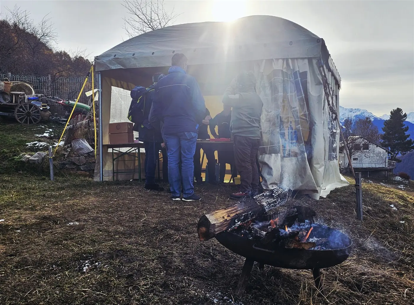 Un gruppo di persone si raduna attorno a una tenda all'aperto, con un fuoco acceso davanti. Sullo sfondo si vedono montagne e alberi. La luce del sole
