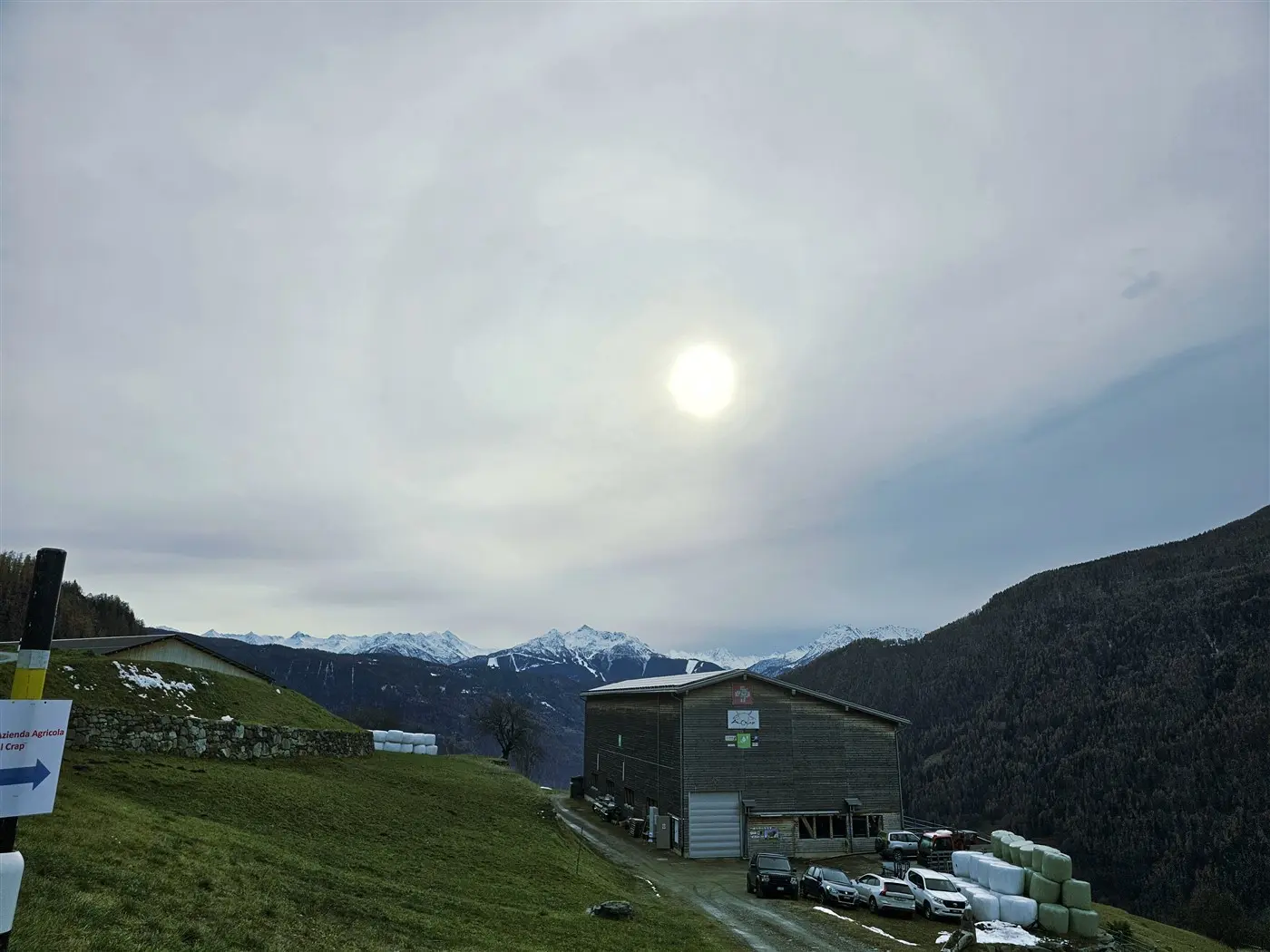 Un paesaggio montano con una cabina di legno, montagne innevate sullo sfondo e un cielo nuvoloso. Il sole è visibile ma velato da nuvole.
