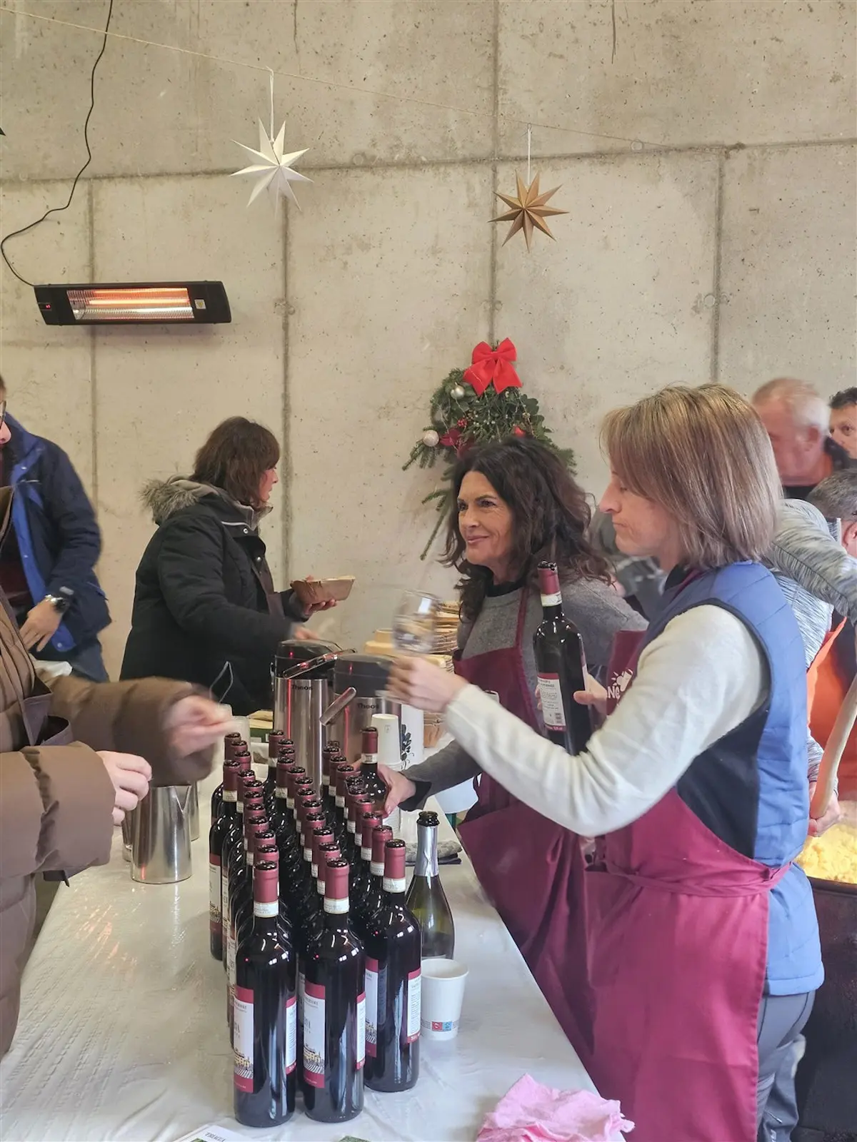 Una folla si riunisce in un ambiente accogliente mentre due donne sorridenti servono vino rosso da una serie di bottiglie su un tavolo. Decorazioni natalizie adornano