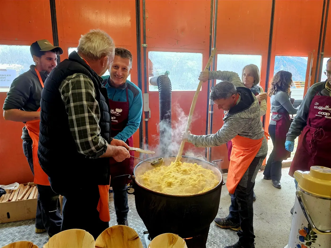 Un gruppo di persone in un laboratorio culinario sta preparando un grande pentolone di polenta, mescolando con cucchiai di legno. Gli partecipanti indossano grembiuli e