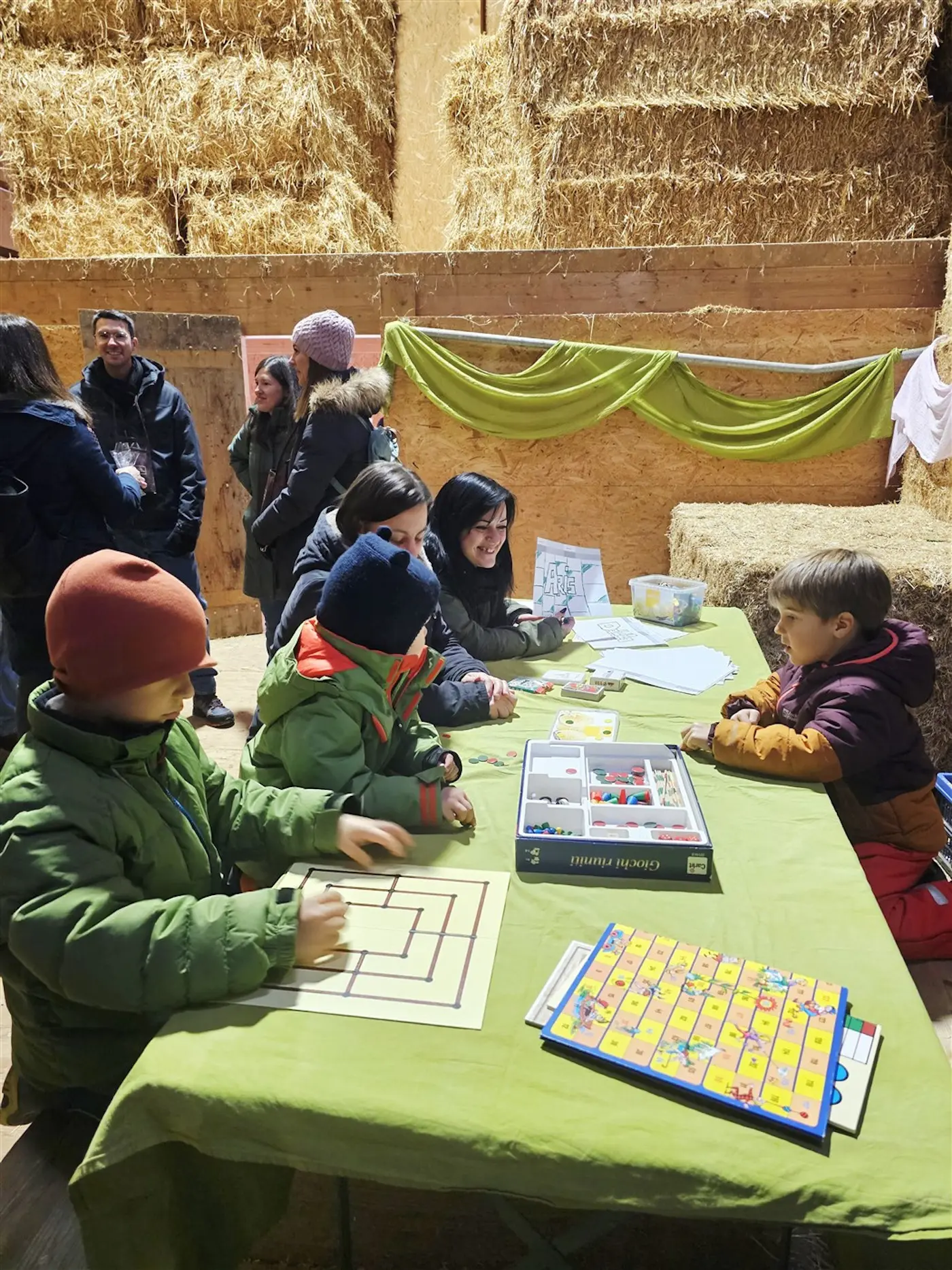 Bambini e adulti partecipano a un'attività ludica all'interno di una stalla, circondati da balle di fieno. Un tavolo verde è coperto di giochi