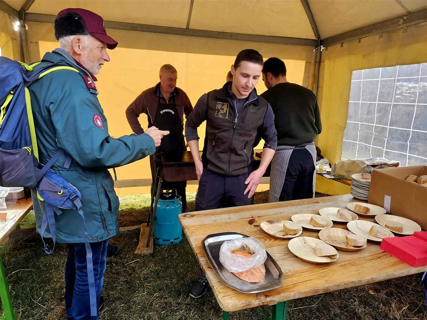 Uomini in un tendone stanno preparando cibo all'aperto. Un uomo in giacca verde parla con un cuoco, mentre altri due stanno cucinando. Ci sono piatti di c