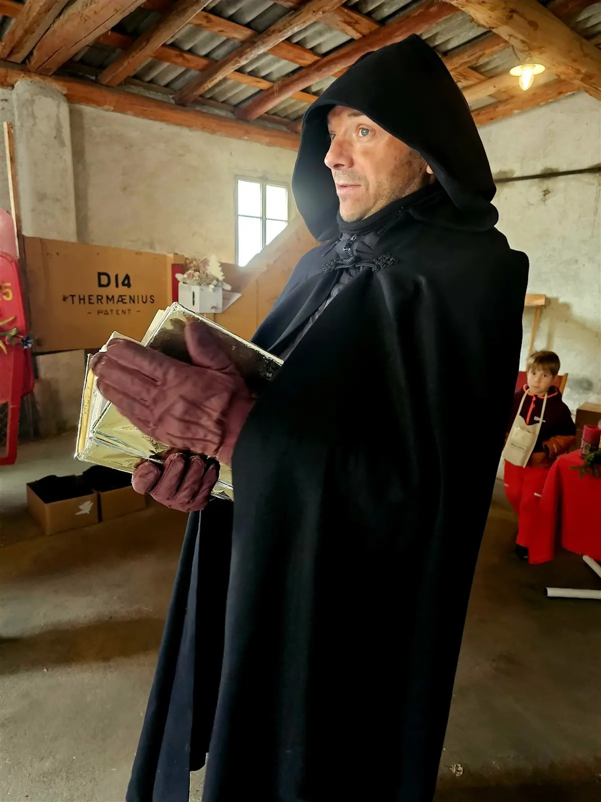 Un uomo in abiti scuri e un cappuccio tiene un libro rilegato in oro. Sguardo serio, si trova in un ambiente rustico con pareti in pietra e