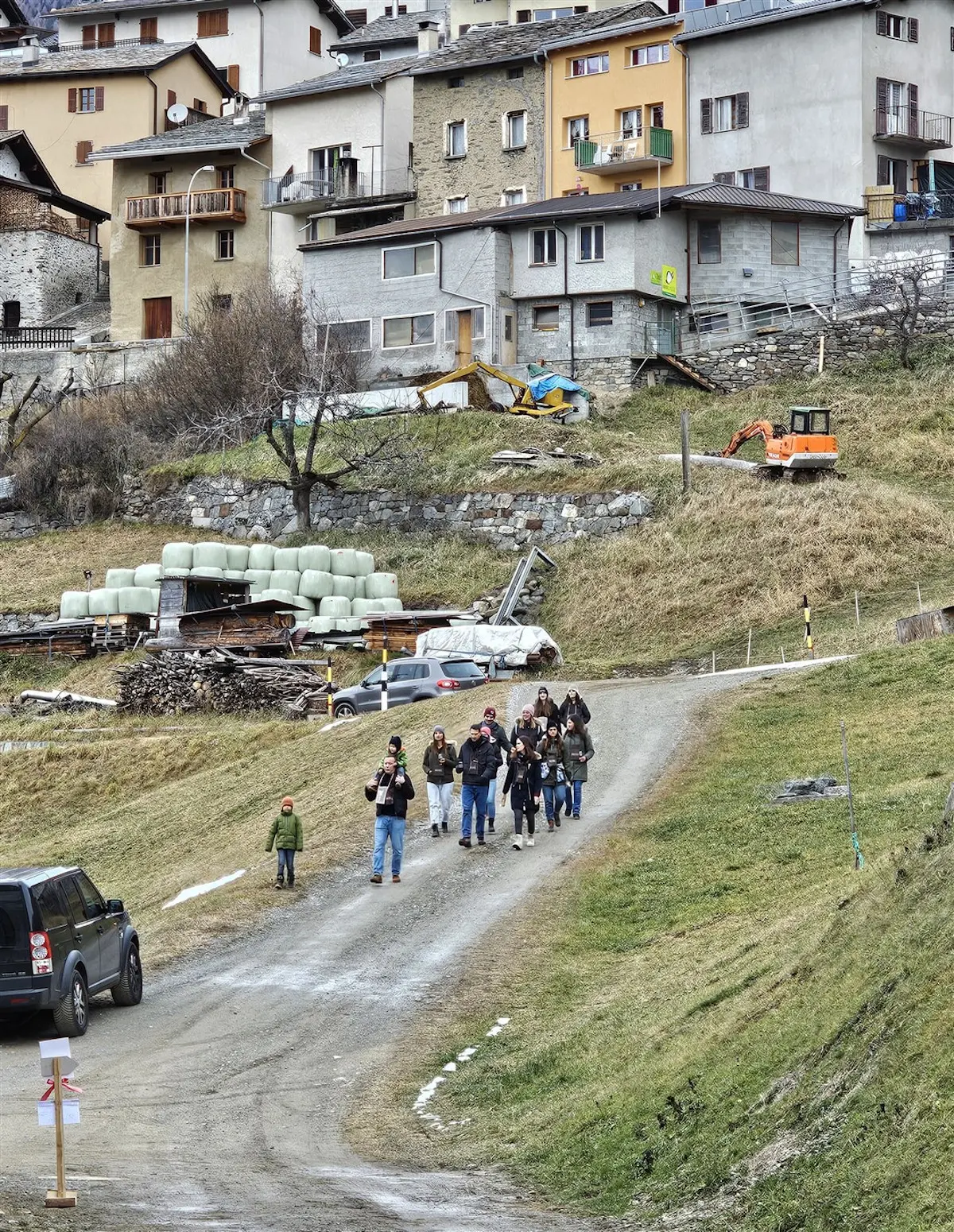 Un gruppo di persone cammina lungo una strada sterrata, circondata da paesaggi verdi e case montane. Alcuni partecipanti portano macchine fotografiche. Sullo