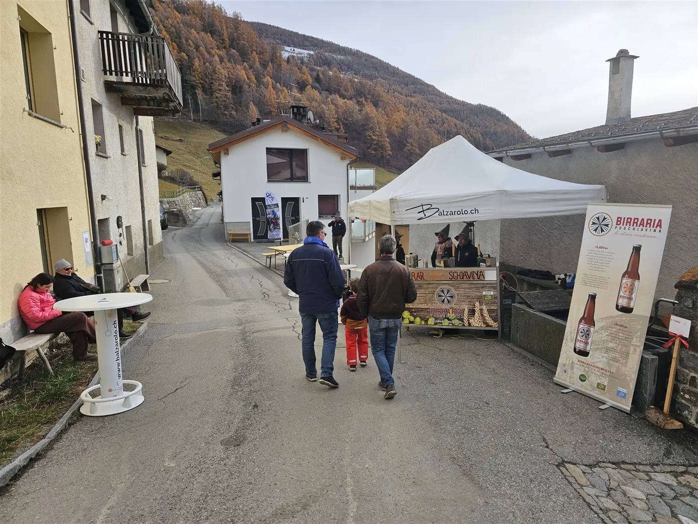Persone che passeggiano lungo una strada di montagna, con un banchetto di birra artigianale e una tenda. Alcuni visitatori sono seduti su panch