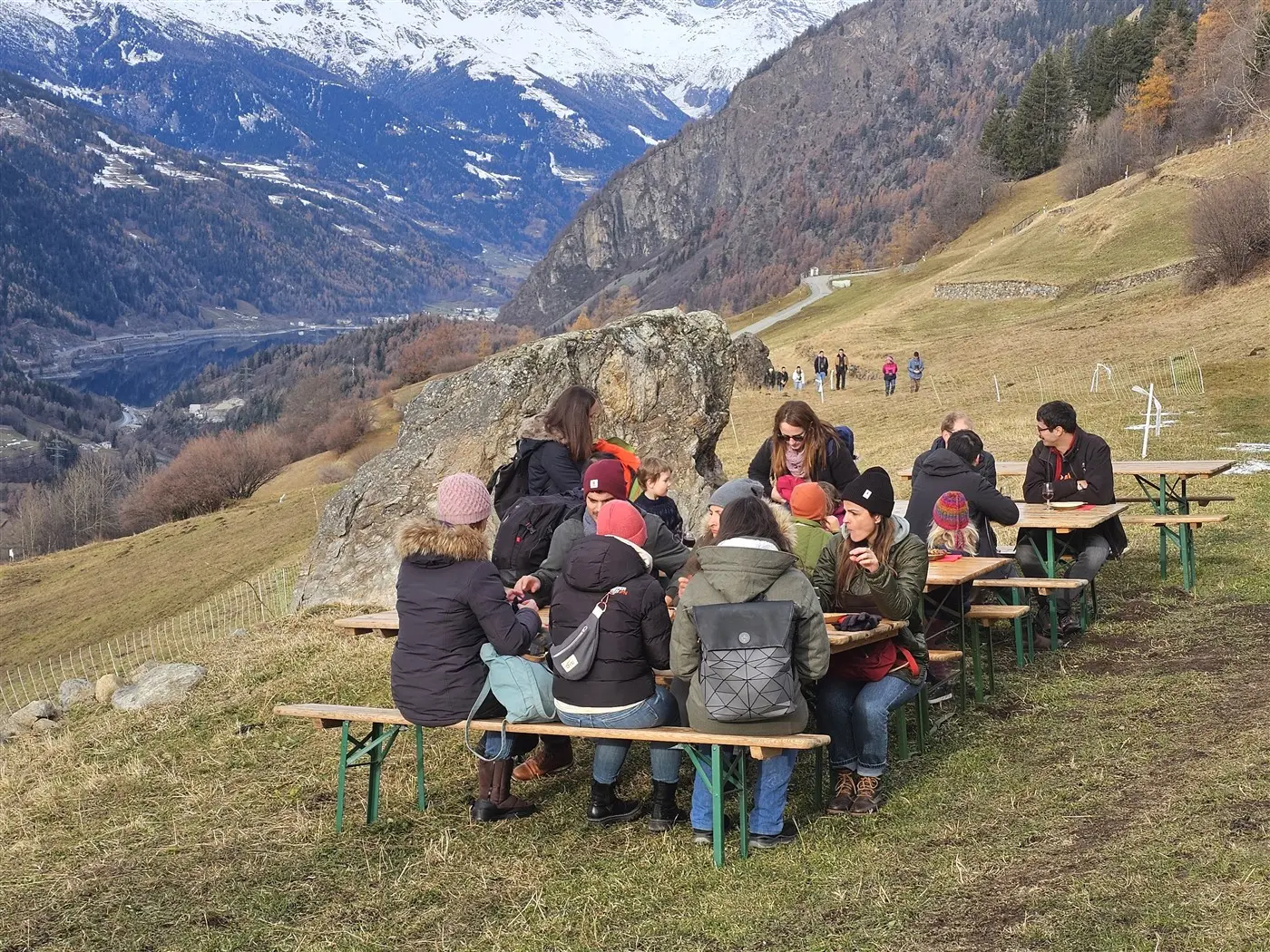 Gruppo di persone sedute a tavoli all'aperto, mentre mangiano e chiacchierano, con montagne innevate e un paesaggio naturale sullo sfondo.