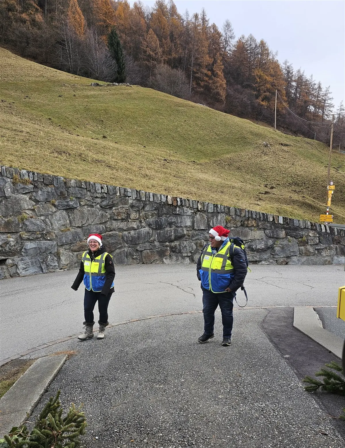Due persone sorridenti indossano giacche fluorescenti mentre si trovano in un'area montuosa. Alle loro teste portano berretti rossi, in un ambiente verde e composto