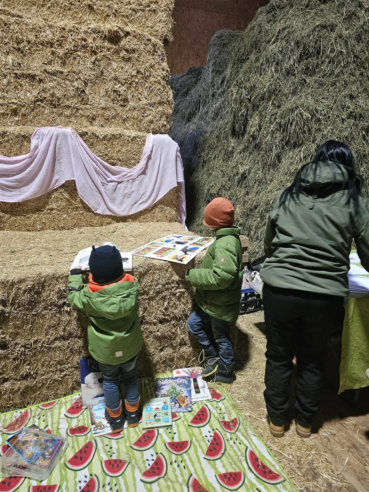 Due bambini in giacche verdi esplorano dei libri su un tavolo accanto a un adulto, con uno sfondo di balle di fieno. Un plaid con motivi di anguria