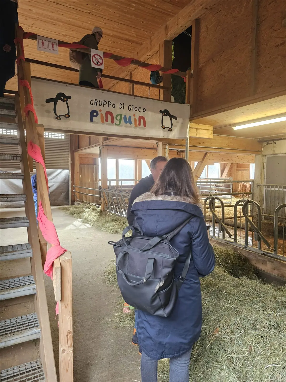 Un gruppo di persone si trova all'interno di una stalla, con una scala che porta a un piano superiore. Un cartello con la scritta "Gruppo Di Gioco Pinguin