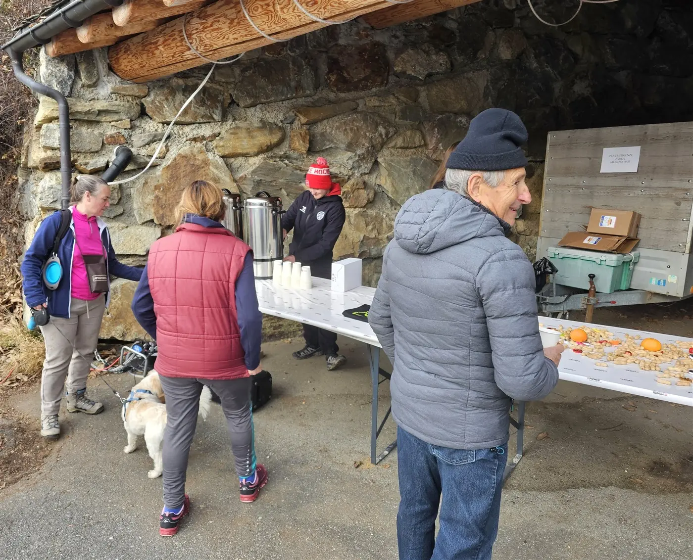 Gruppo di persone all'aperto, vicino a un tavolo con cibo e bevande. Due persone sono in piedi, una con un cappello, e un'altra è voltata verso di