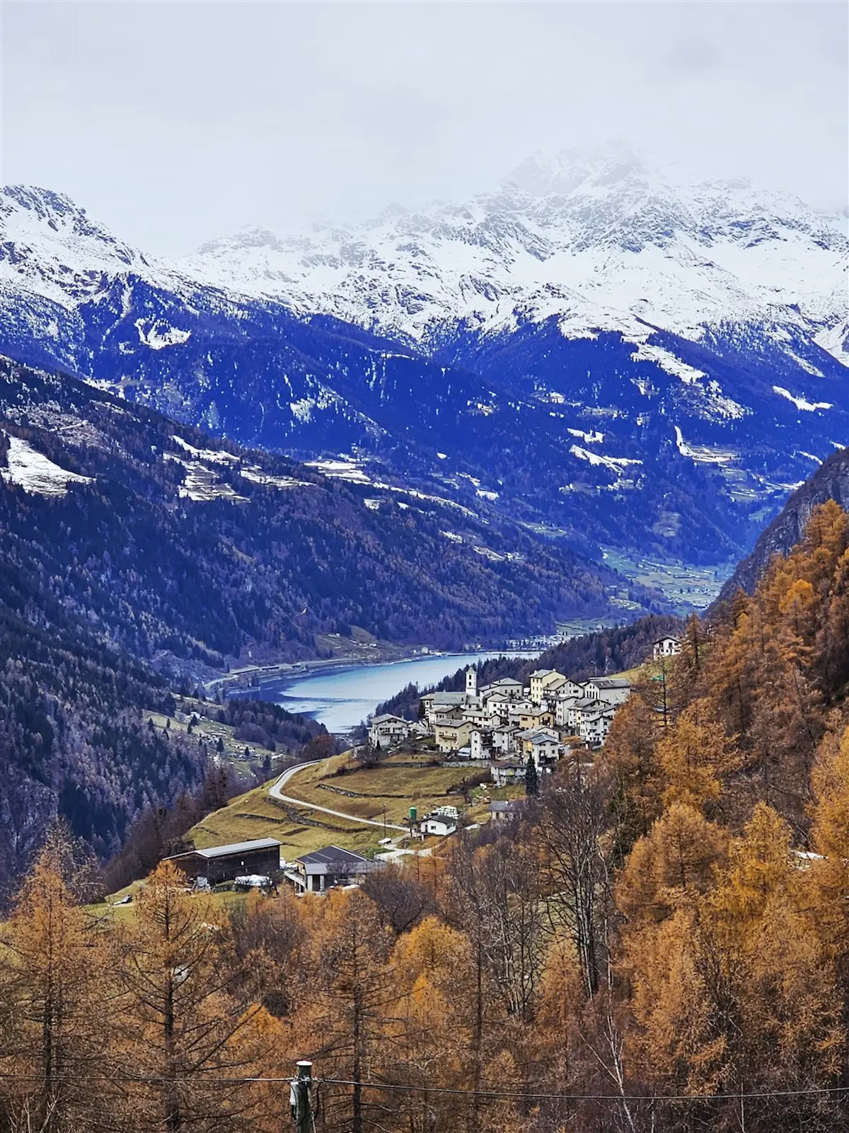 Panorama montano con un villaggio circondato da alberi dorati e cime innevate. Un lago lucente è visibile in lontananza, mentre le nuvole coprono